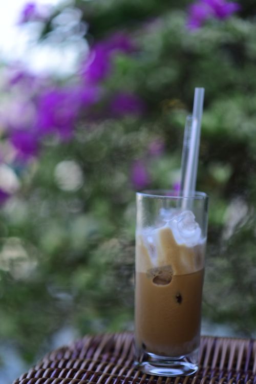Free A tall glass of iced coffee with milk on a woven table, set against a blurred floral backdrop. Stock Photo