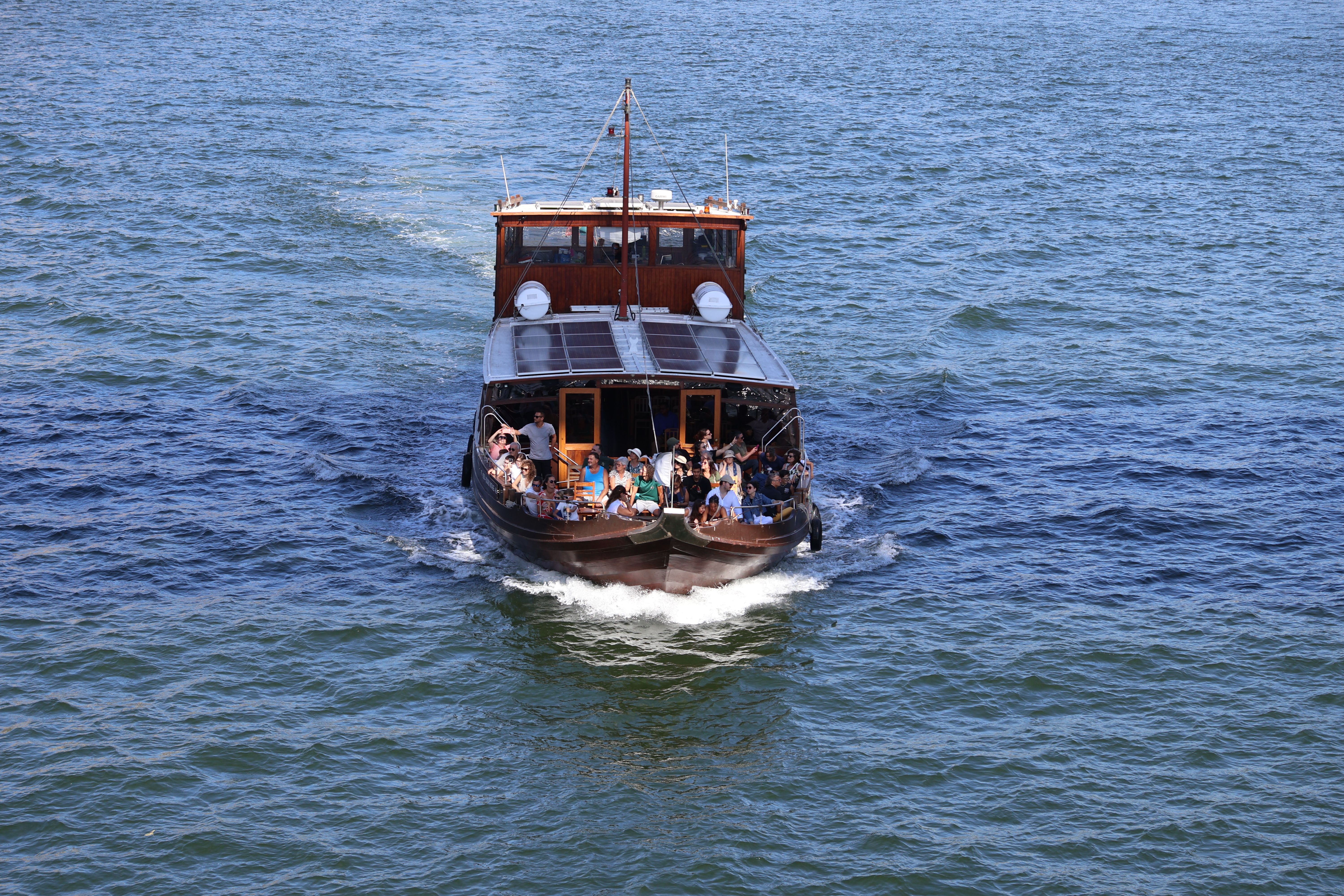 Small wooden boat with several people sailing across blue water, leaving a white wake.