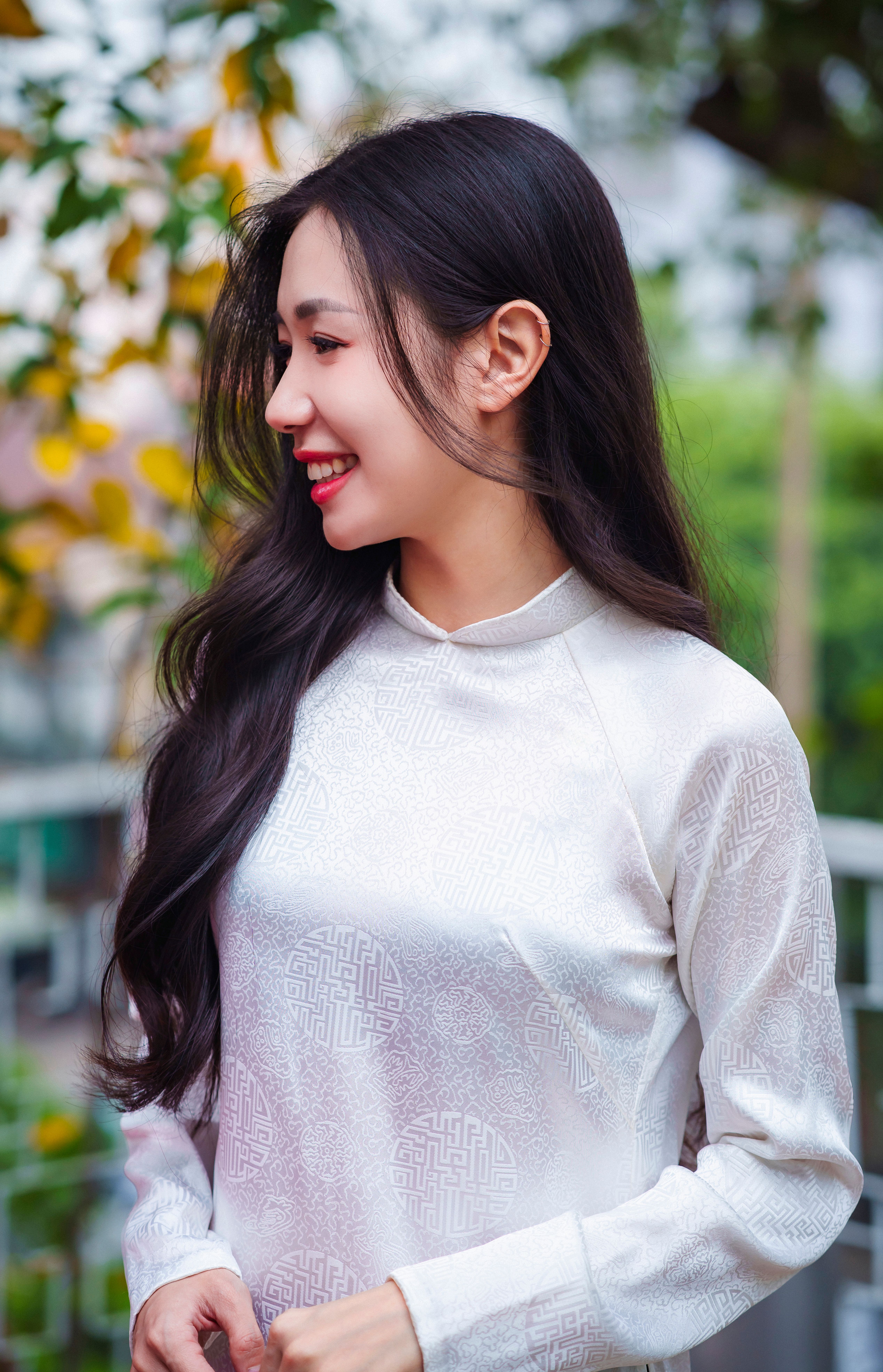 A woman in an elegant white ao dai stands smiling in an outdoor setting, reflecting Vietnamese culture.