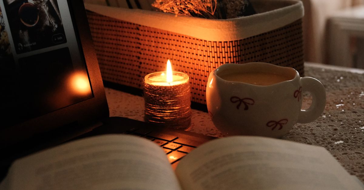 A messy desk with books, a laptop, and a steaming mug, lit by a desk lamp in a dark room