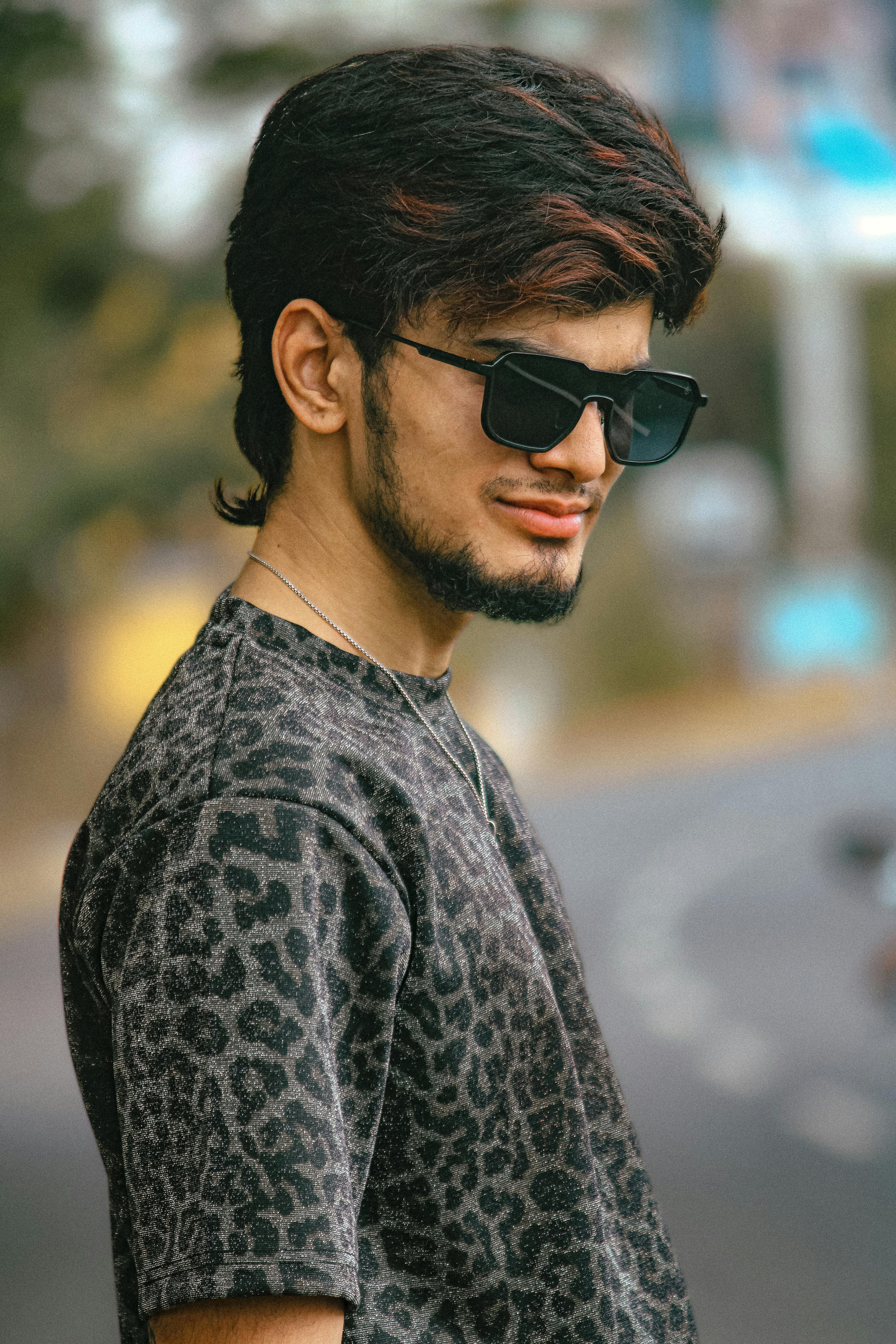 A fashionable young man wearing sunglasses and a patterned shirt poses confidently outdoors.