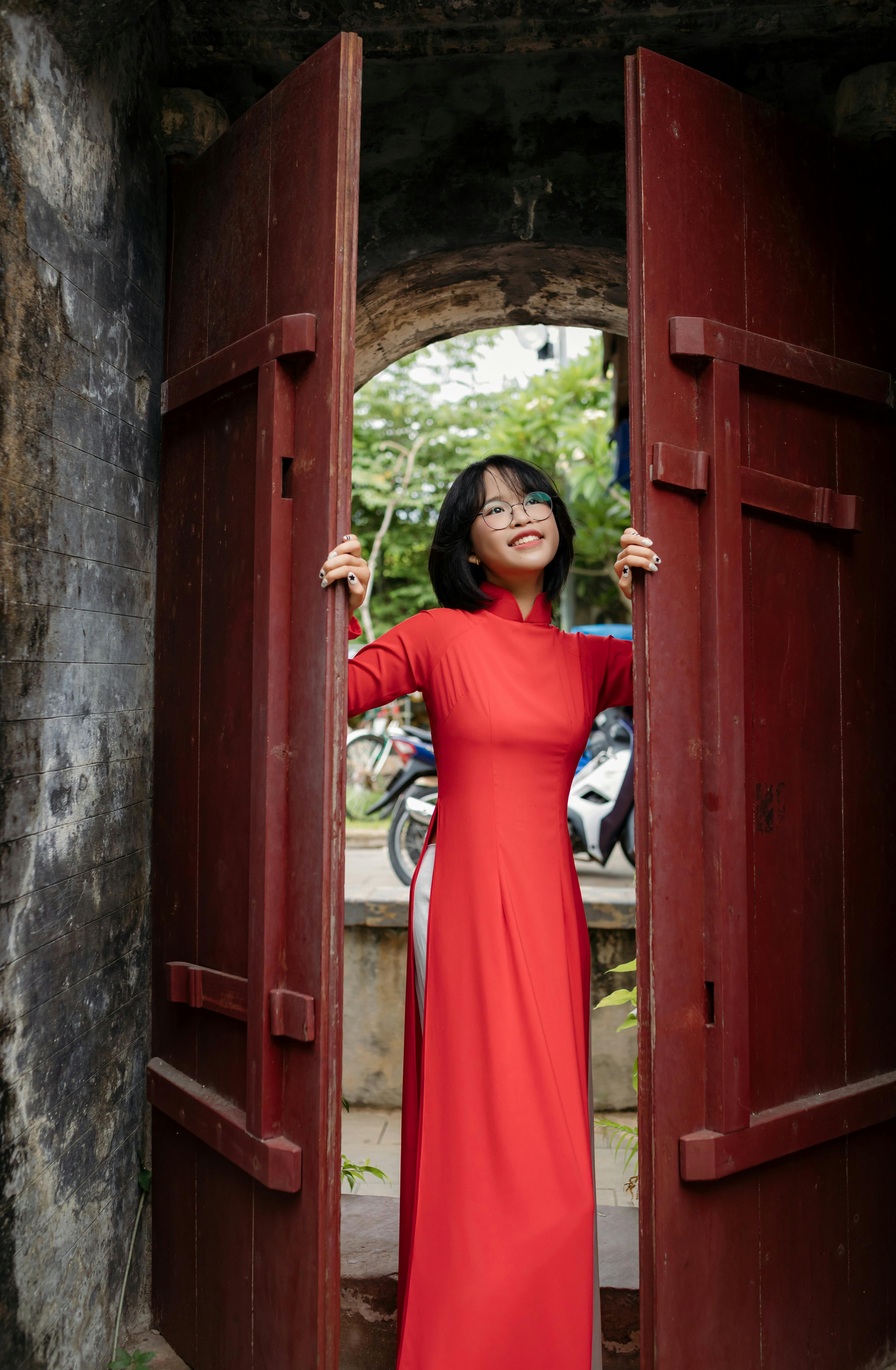 Smiling woman in red ao dai posing at historic wooden gate in Hoi An, Vietnam.