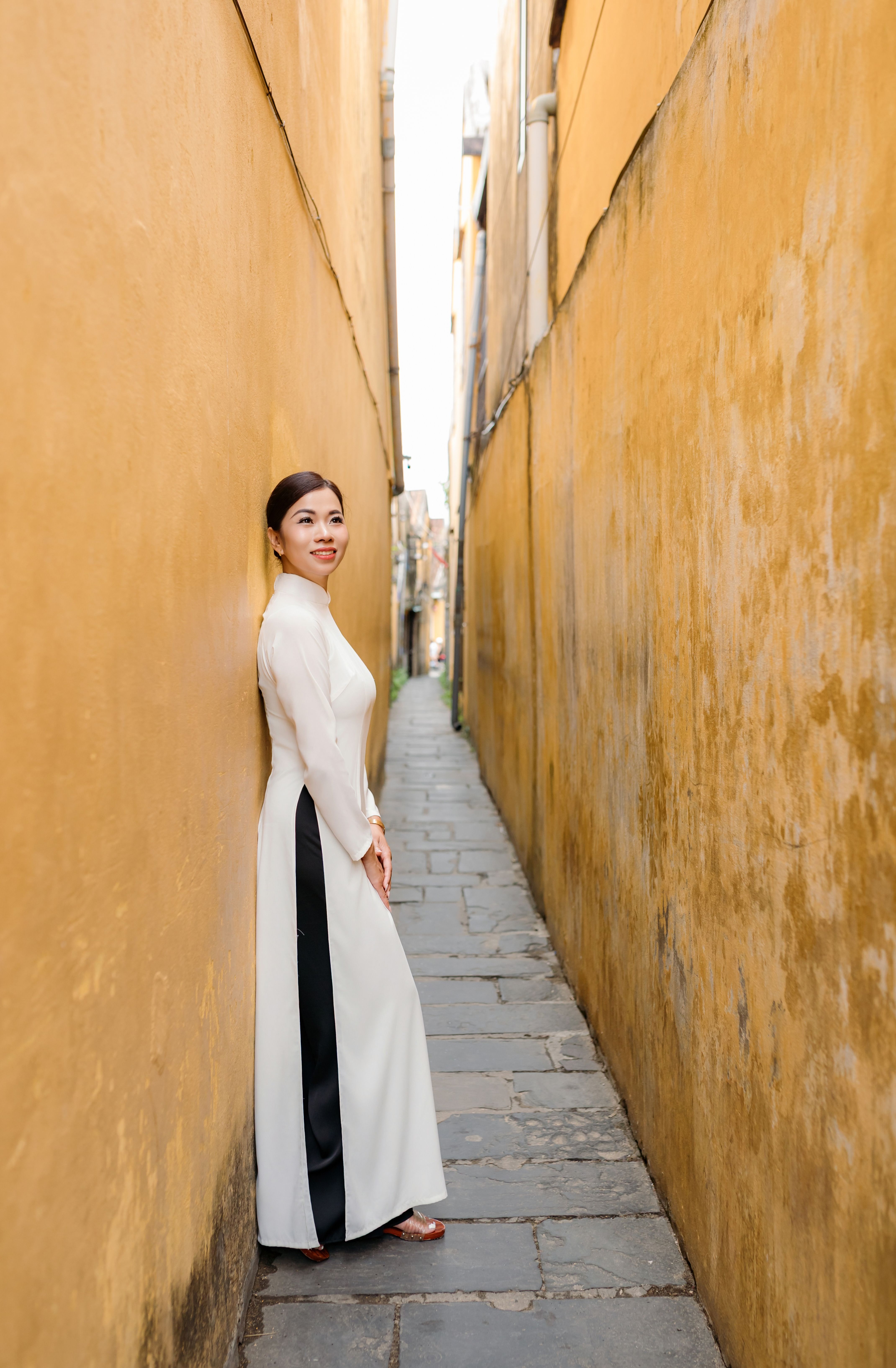 A woman in a traditional Ao Dai stands in a narrow, historic alley in charming Hoi An, Vietnam.