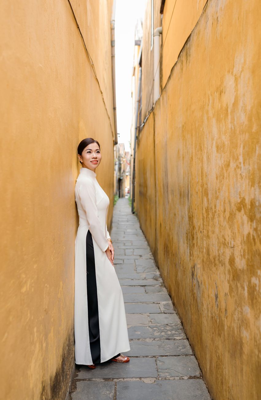 A woman in a traditional Ao Dai stands in a narrow, historic alley in charming Hoi An, Vietnam.