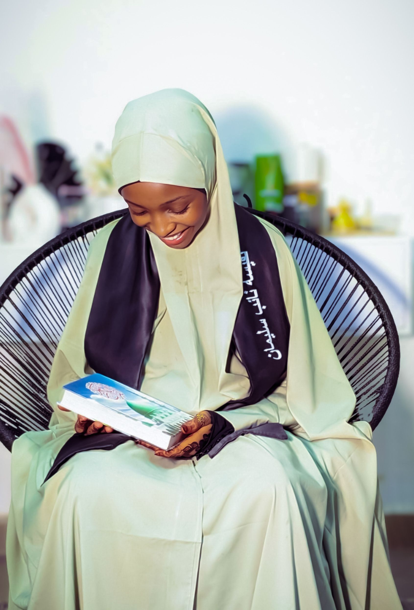 A young woman in a hijab smiling while reading a book indoors, conveying warmth.