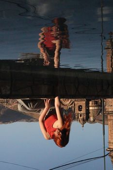 Reflection of a woman in red tank top by a historic building in water.