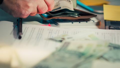 Close-up of hands counting cash next to a wallet and documents on a desk.