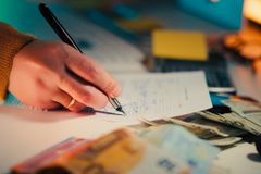 Close-up of a hand writing on documents with euro notes scattered on a desk, focusing on finance and business.
