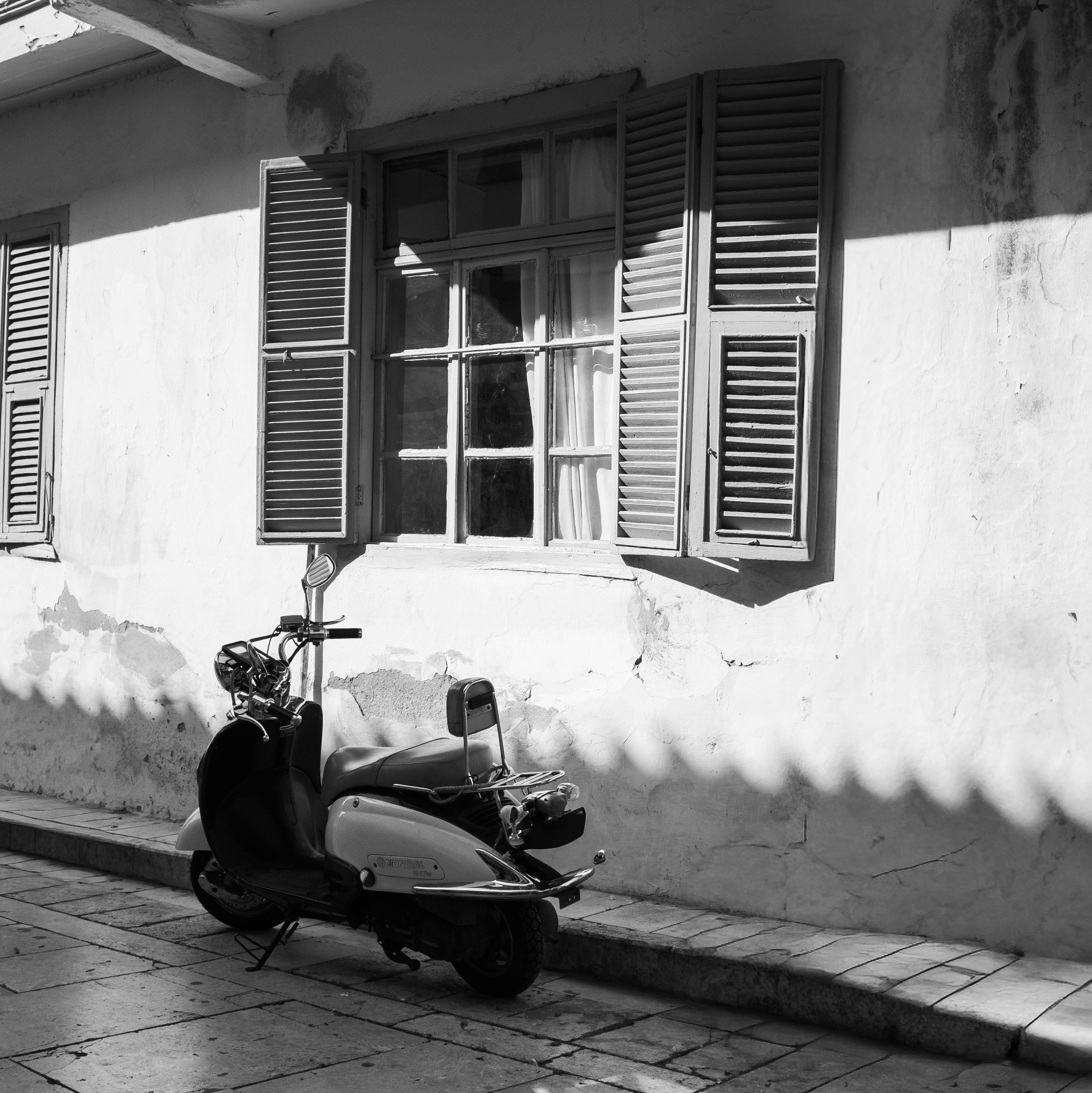 Black and white image of a scooter parked by a sunlit window with shutters casting shadows.
