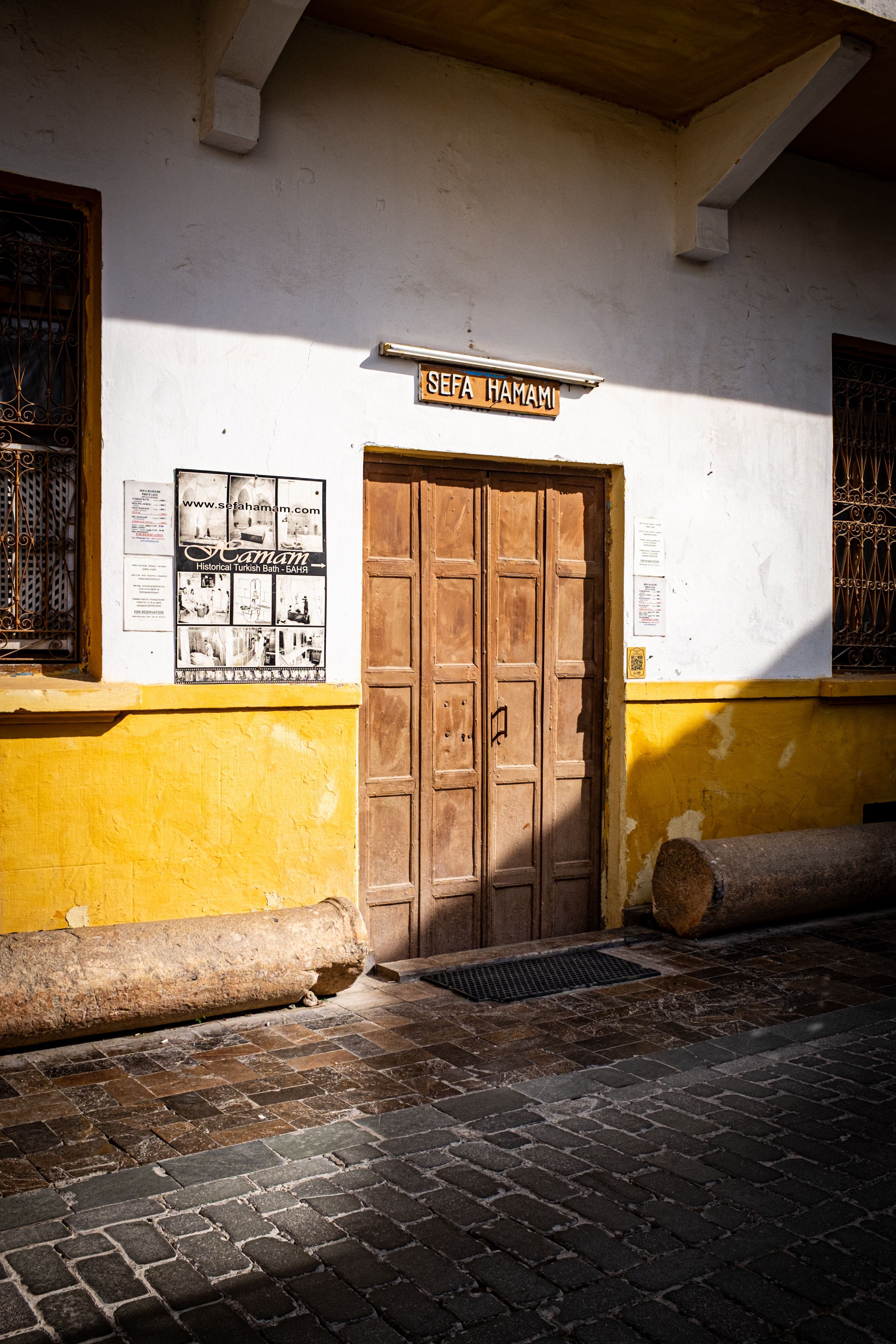 Front view of Sefa Hamam entrance with rustic wooden door and sunlight on cobblestone street.