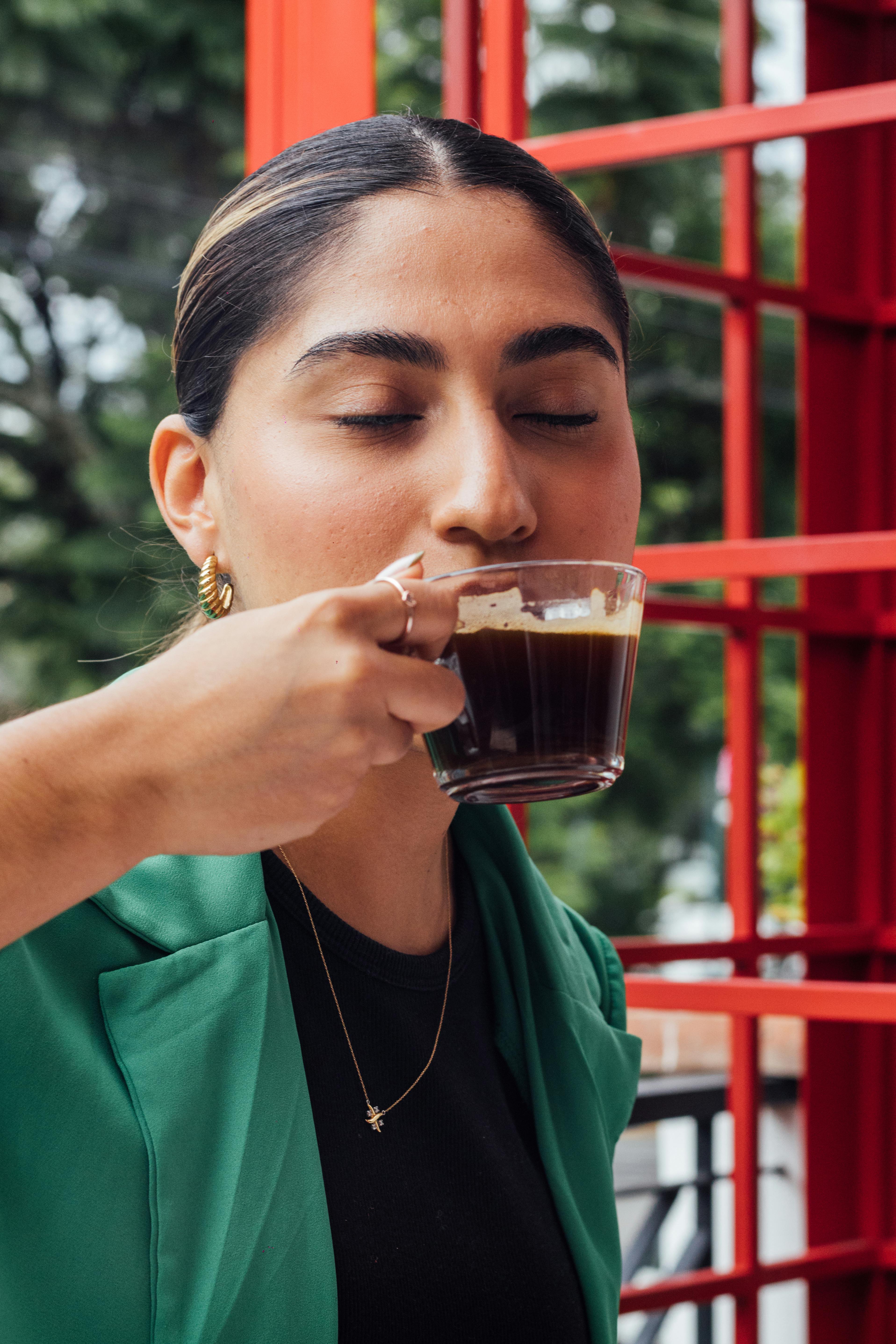 Adult woman savoring a hot espresso outdoors, eyes closed, relaxing moment.