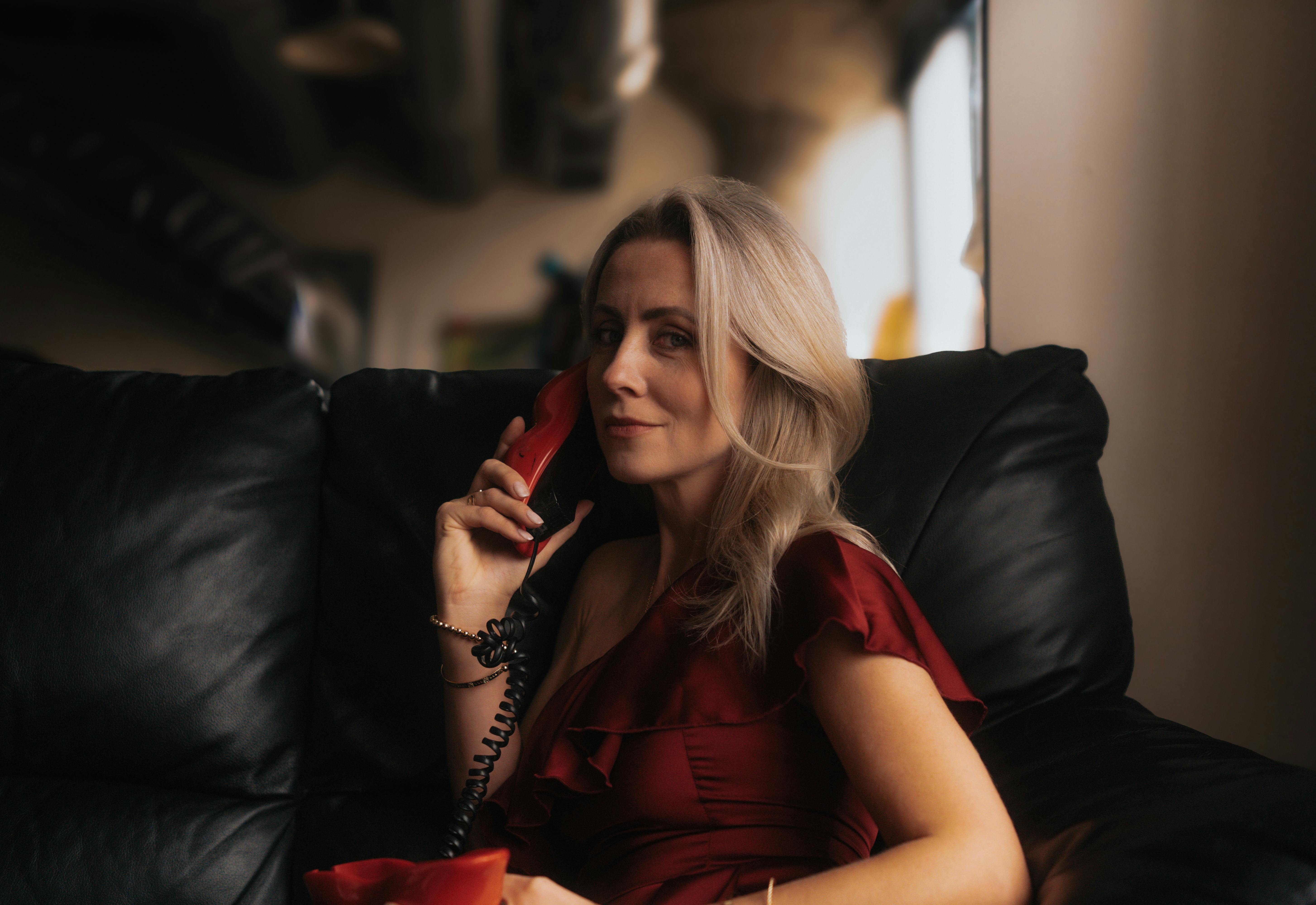 Miễn phí Người phụ nữ sành điệu trong chiếc váy đỏ đang sử dụng điện thoại cổ điển trên chiếc ghế sofa da. Ảnh lưu trữ