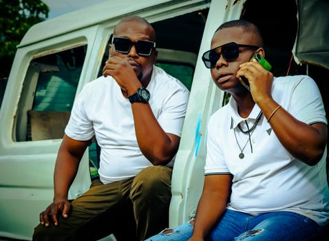 Two men in white t-shirts and sunglasses pose by an old car in South Africa.