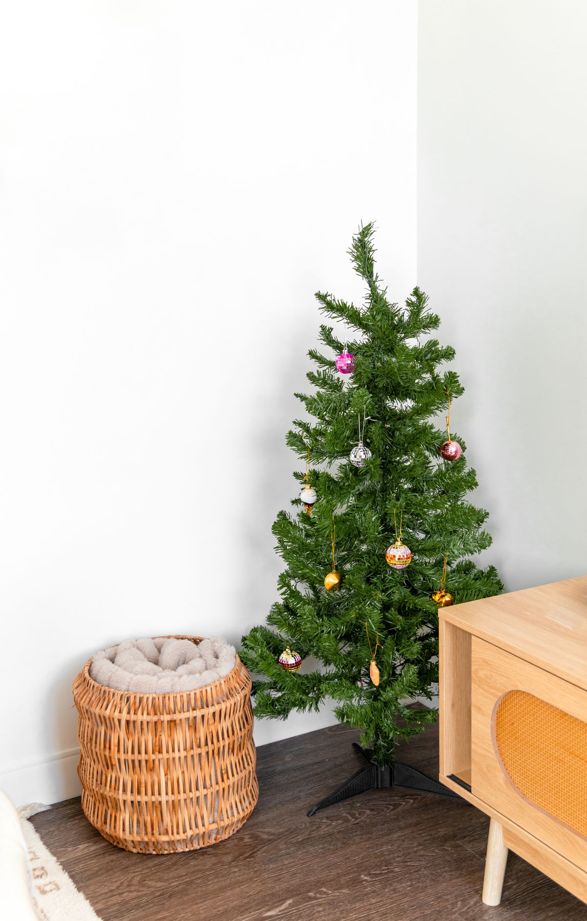 Small apartment Christmas decor: a narrow three-foot Christmas tree beside a woven basket and a wooden box against a white wall