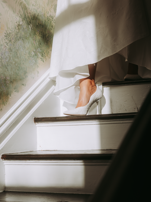 Free Bride gracefully walks down sunlit stairs, showcasing elegant white heels. Stock Photo