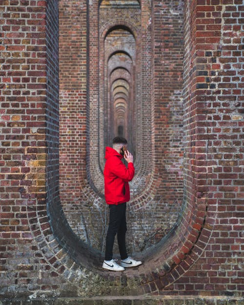 Man in Red Jacket and Black Pants Standing on Red Brick Wall