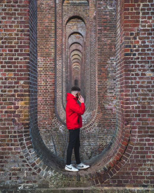 Man in a vibrant red jacket stands between repeating brick arches, creating a depth effect.