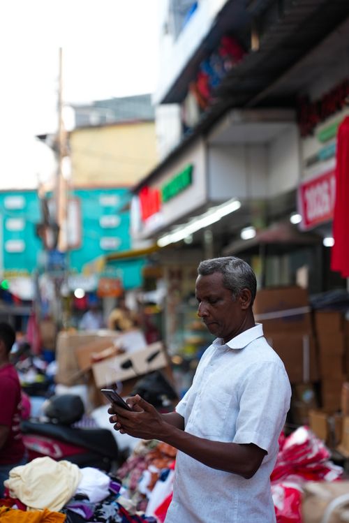 Free A middle-aged man wearing a white shirt uses his smartphone in a vibrant outdoor market. Stock Photo