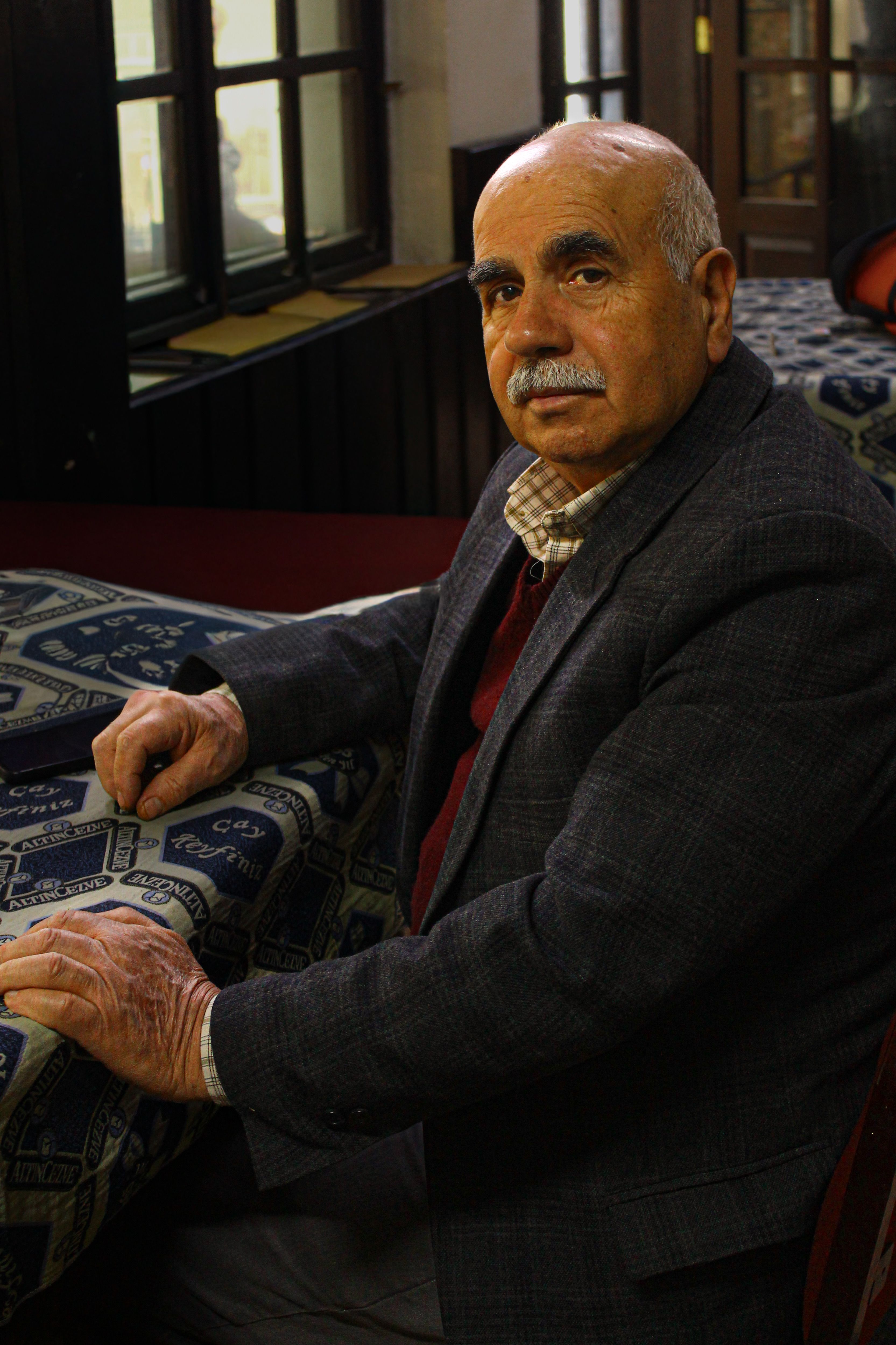 Elderly man seated indoors, wearing a smart-casual outfit. Thoughtful expression.