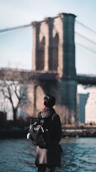 A person with a backpack stands near Brooklyn Bridge, New York City, capturing the essence of travel. by Rick Han