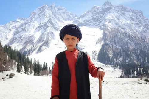 Free A young boy in traditional clothing stands before snow-covered mountains in Sonamarg, Kashmir. Stock Photo
