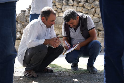Two men squatting and analyzing paperwork outdoors in a sunny setting.