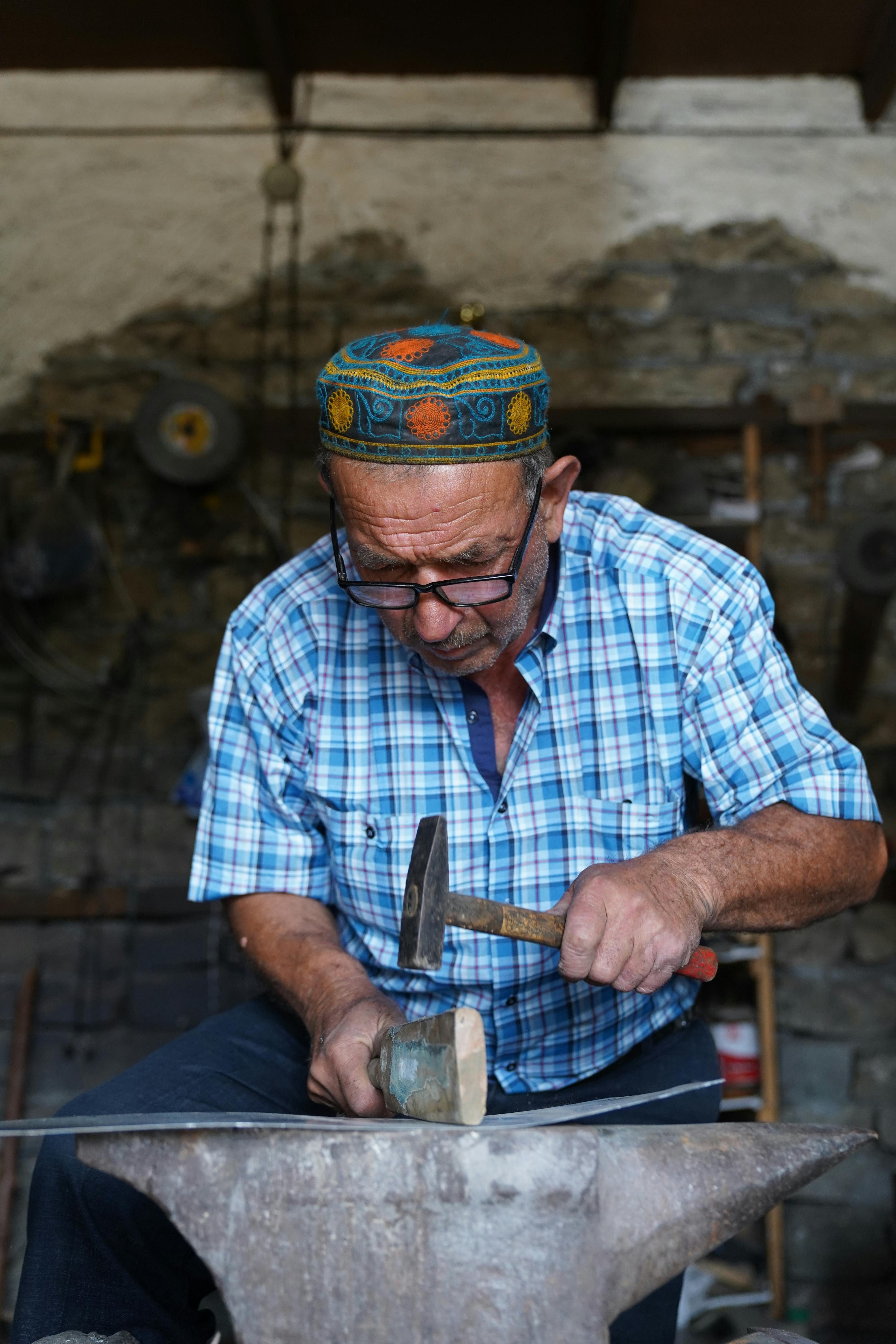 Elderly blacksmith shaping metal on anvil in a traditional workshop setting.