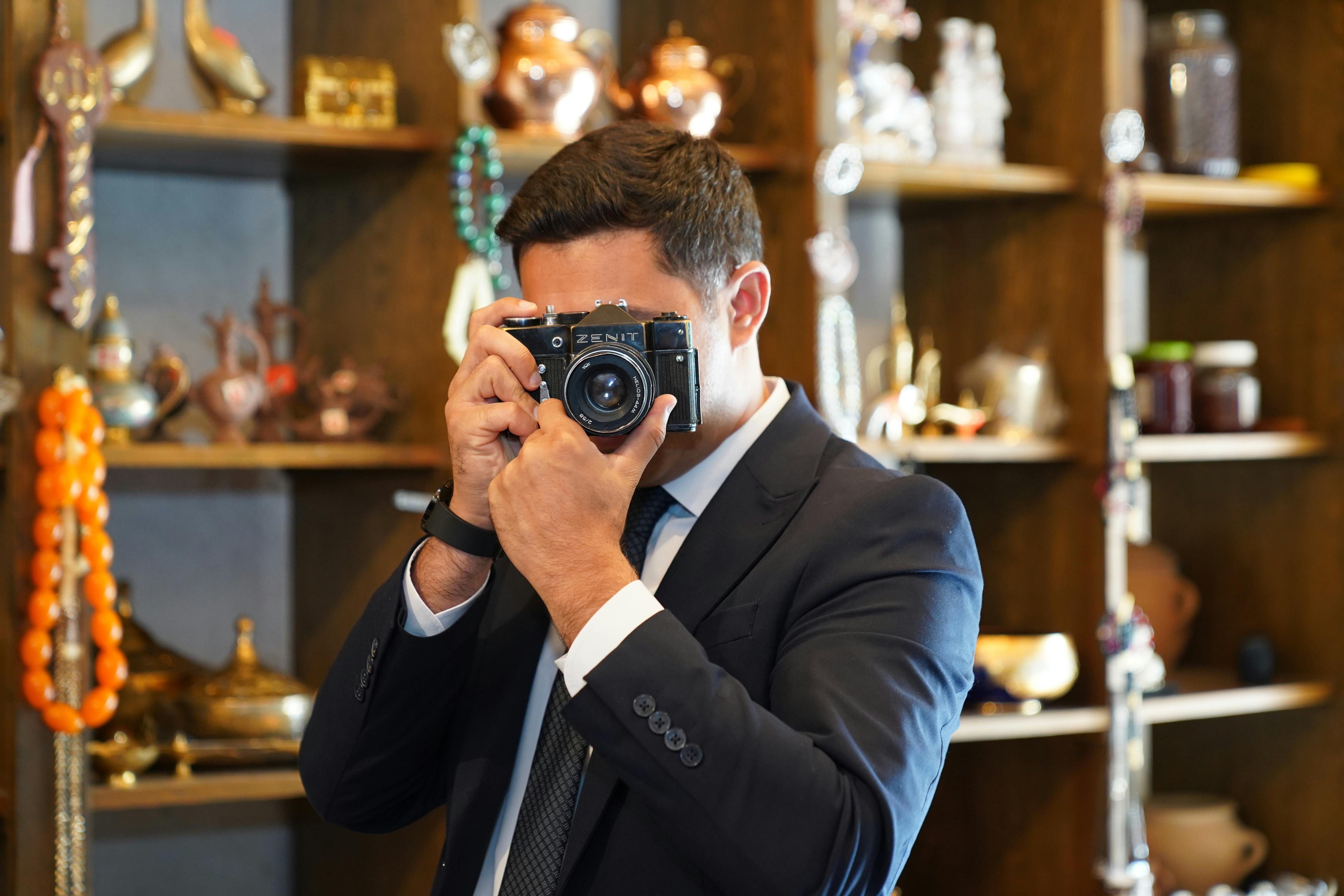 A man in a suit taking a picture with a vintage camera in a store with various artifacts.