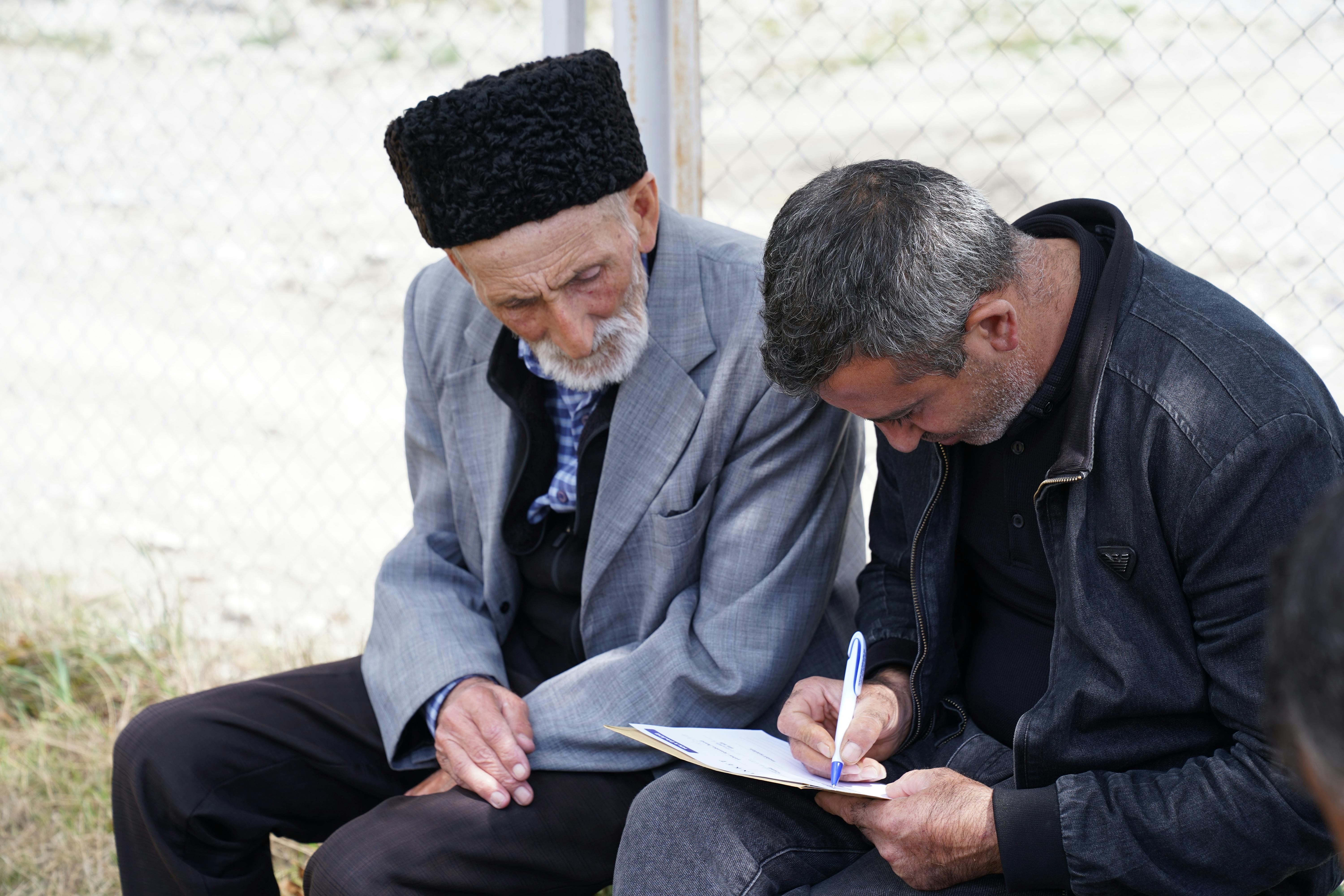 A man helps an elderly person fill out a form while sitting outdoors.