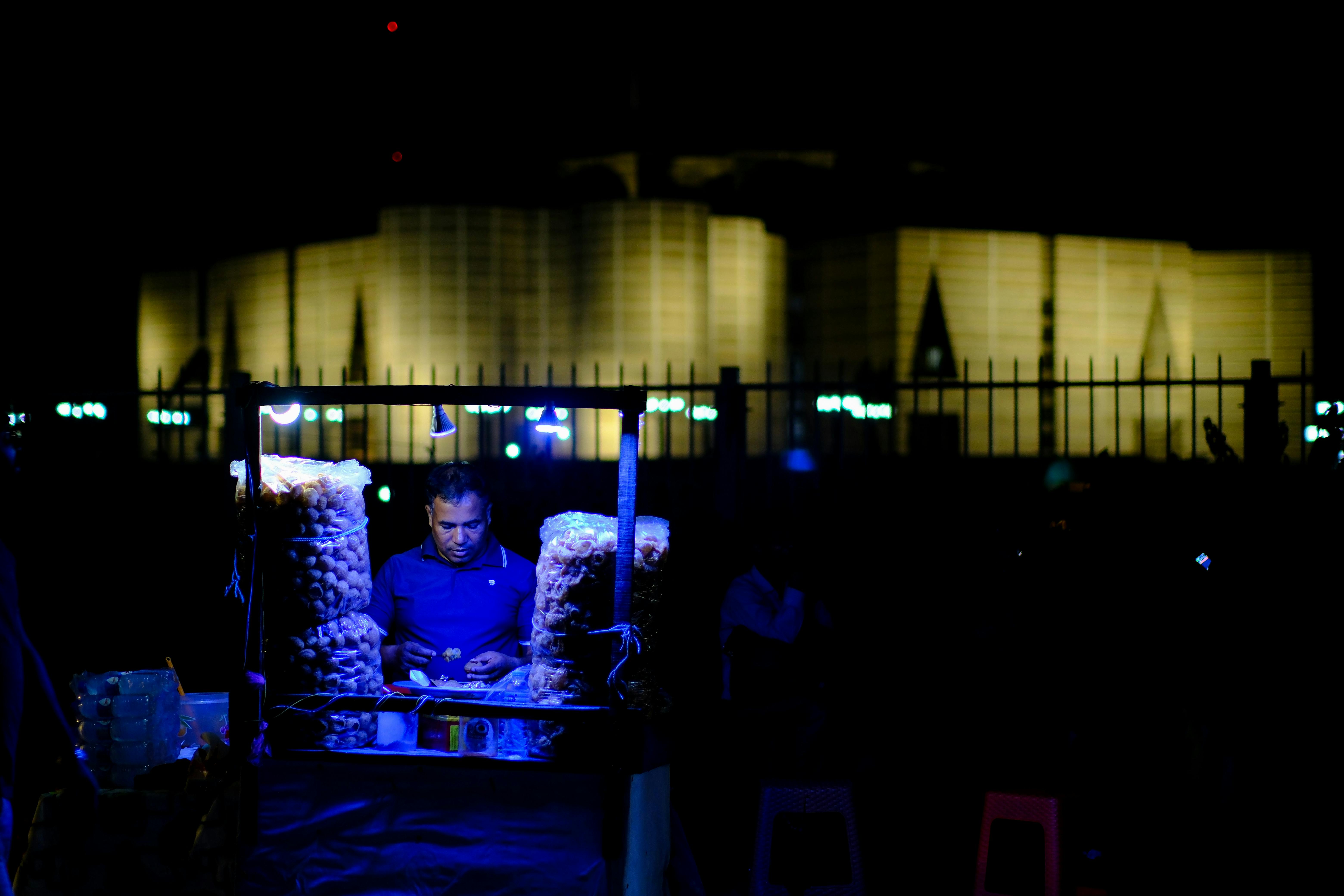 Street vendor selling snacks at night in front of iconic structure in Dhaka, Bangladesh.