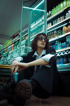 Young person with curly hair and sunglasses sitting in a supermarket aisle.
