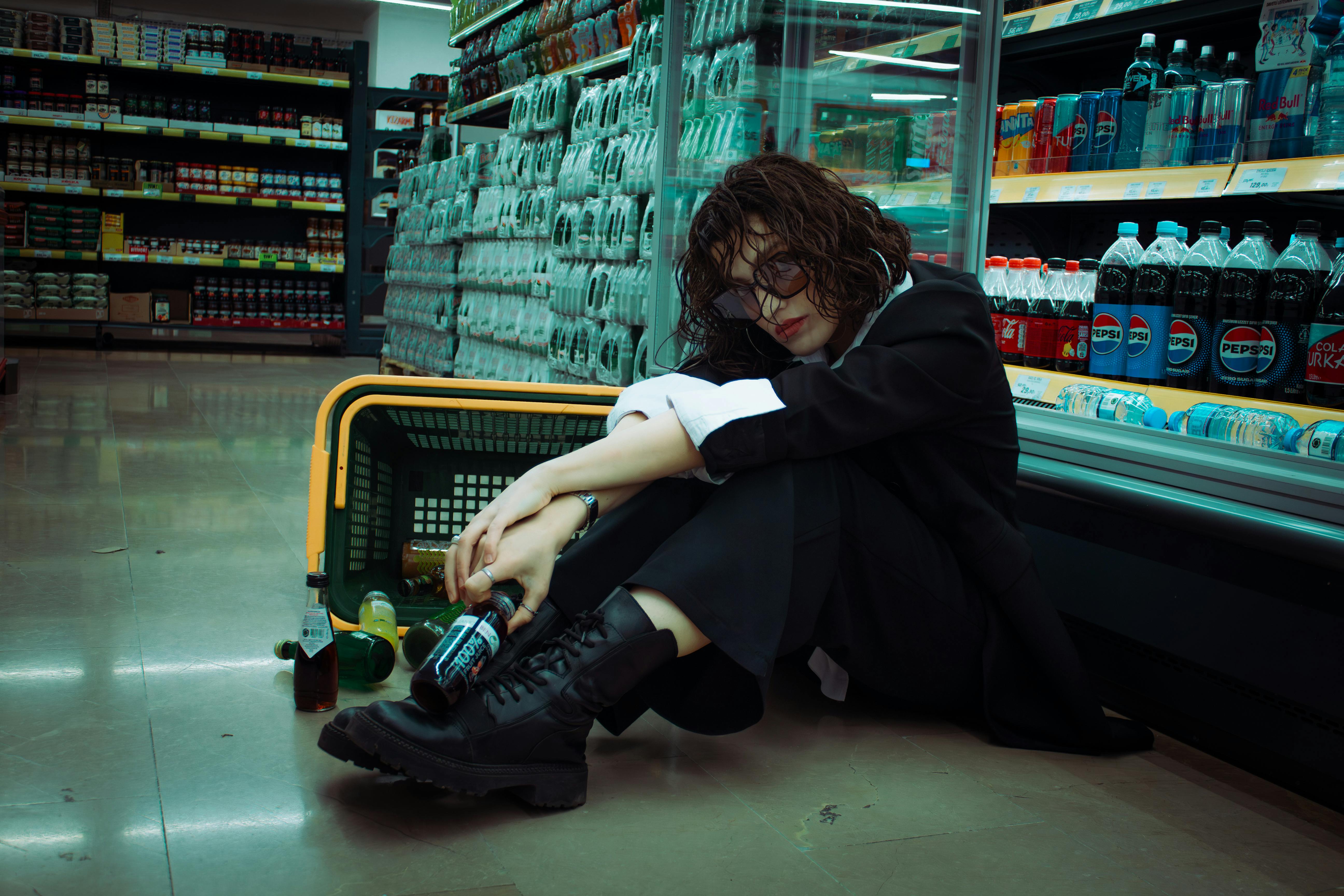 A young adult sits in a supermarket, surrounded by drink bottles and a shopping basket.