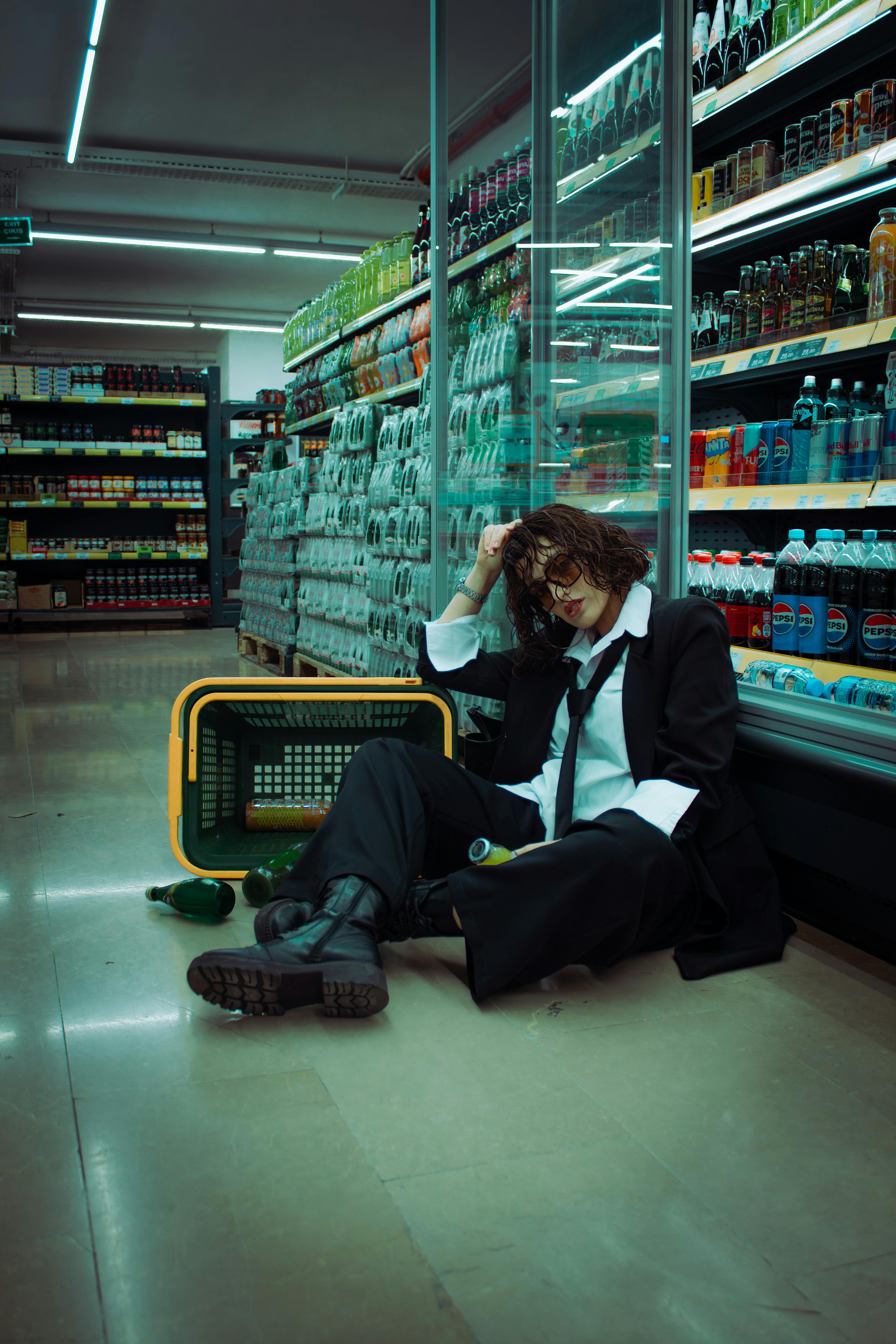 Person sits on supermarket floor, surrounded by drink bottles, conveying a mood of stress and fatigue.