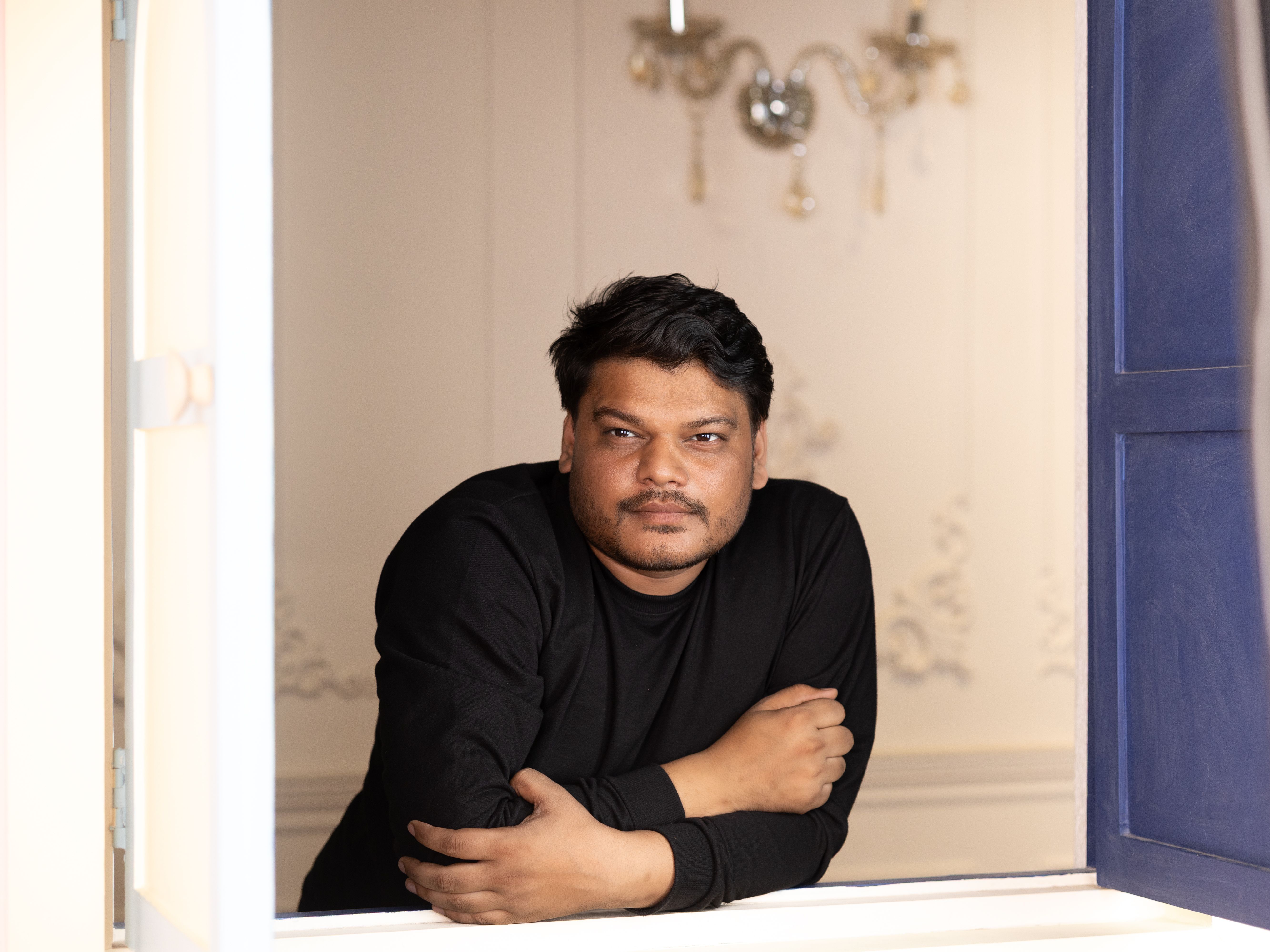 A relaxed man in a black shirt leaning on a window ledge indoors.