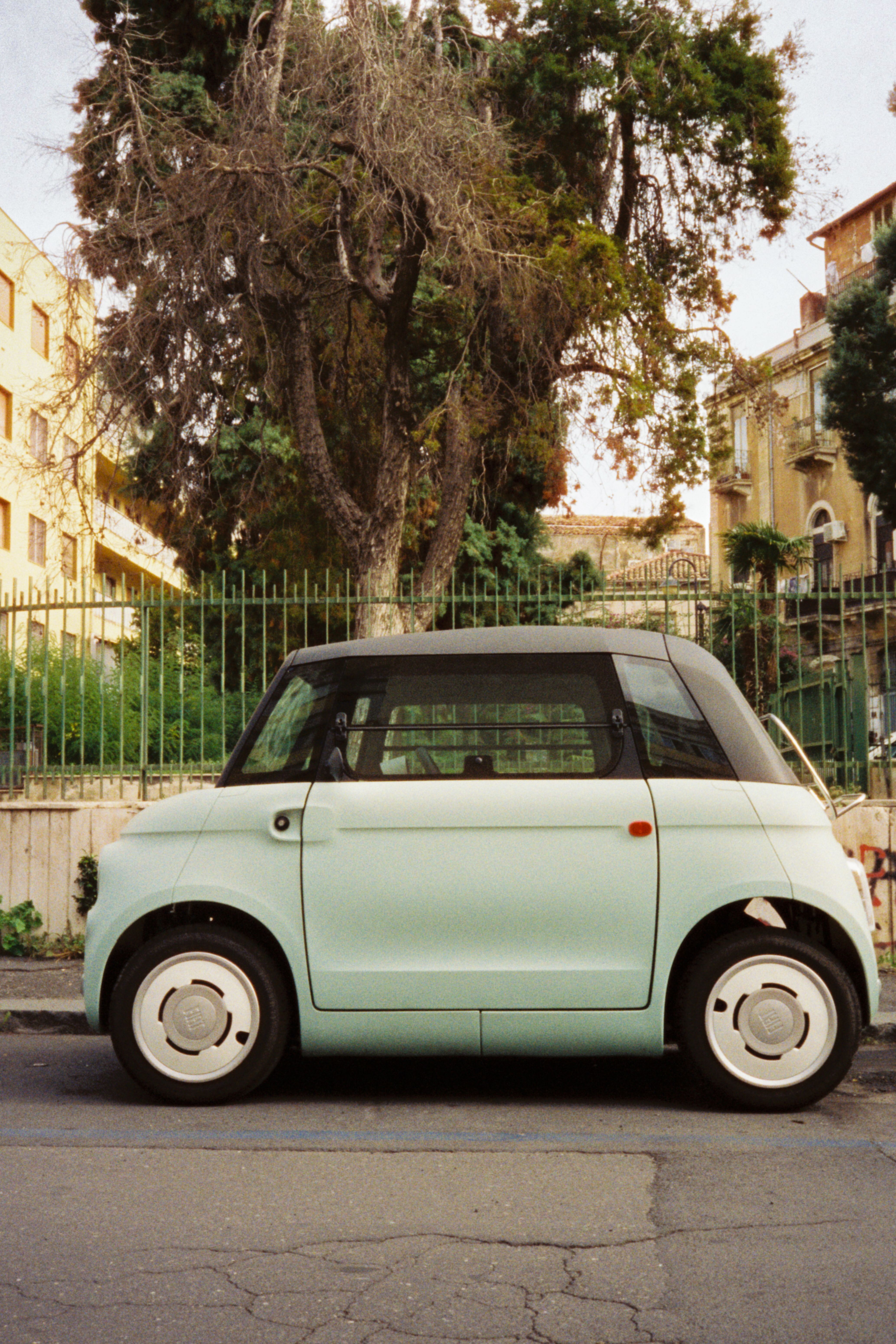 A vintage compact car parked on a street in Sicily, Italy, with lush greenery in the background.