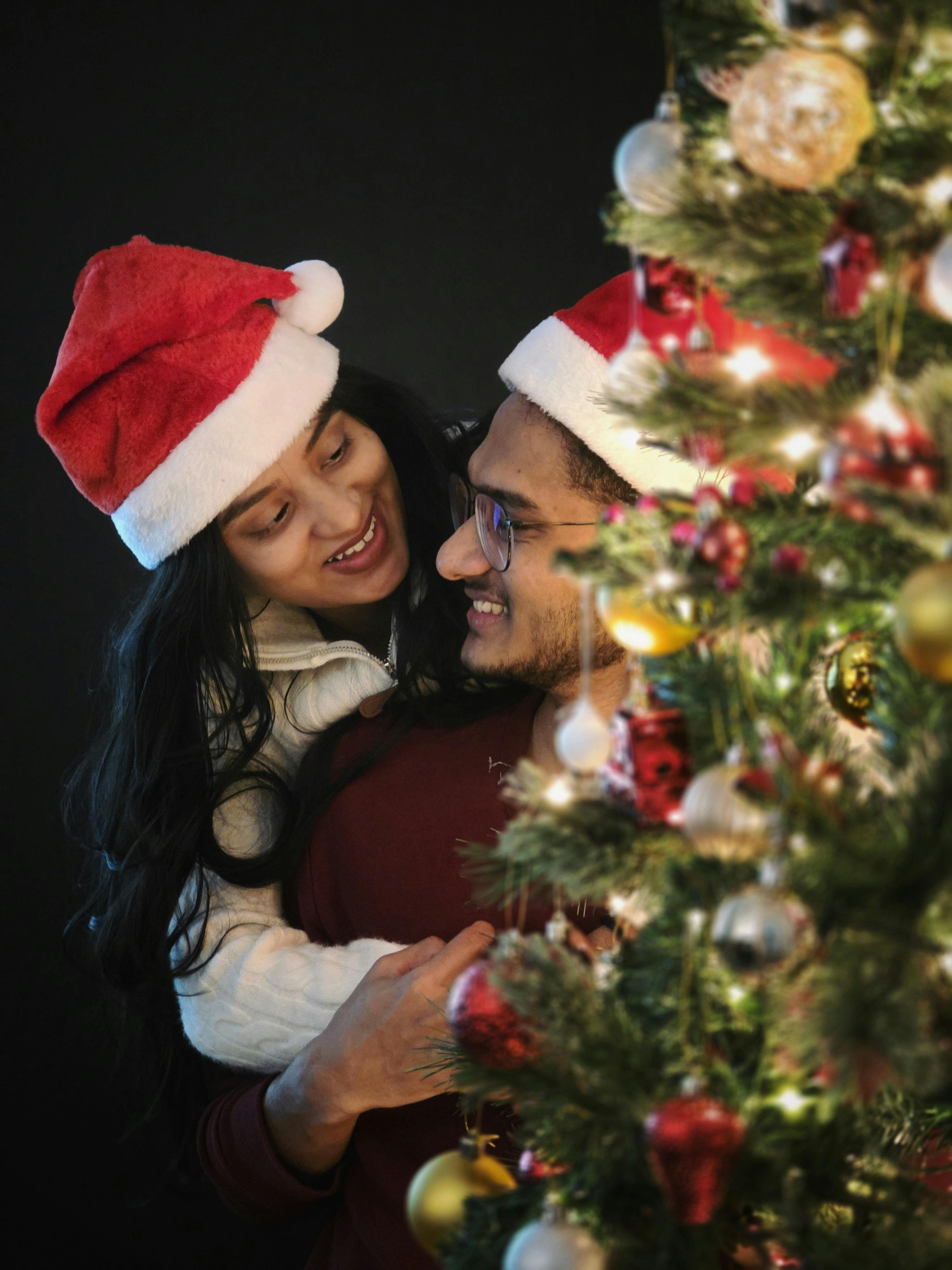 Smiling couple in Santa hats by a Christmas tree with decorations.
