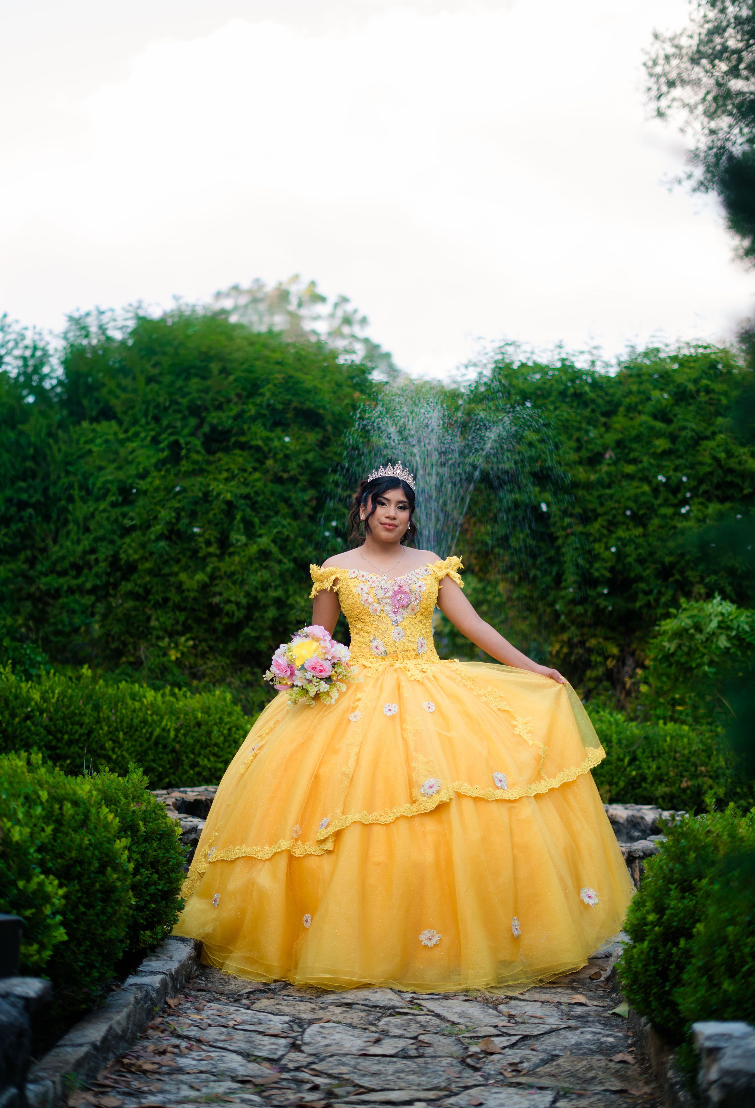 A young girl in a yellow quinceañera dress poses in a lush garden.