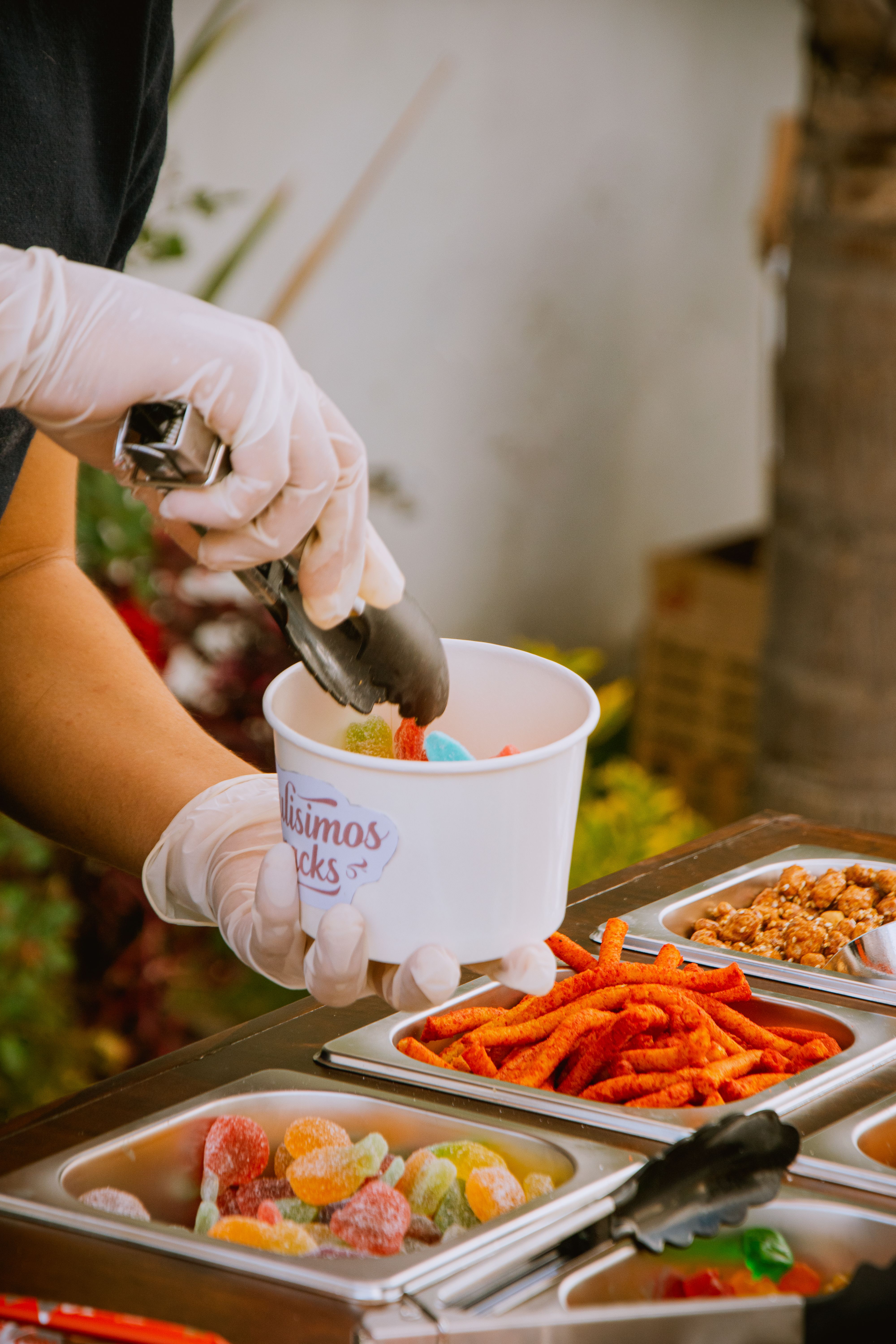 Gloved vendor serving assorted colorful snacks into a cup at an outdoor market.