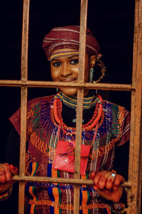 Free A woman in traditional African attire poses cheerfully behind bars, showcasing cultural jewelry. Stock Photo