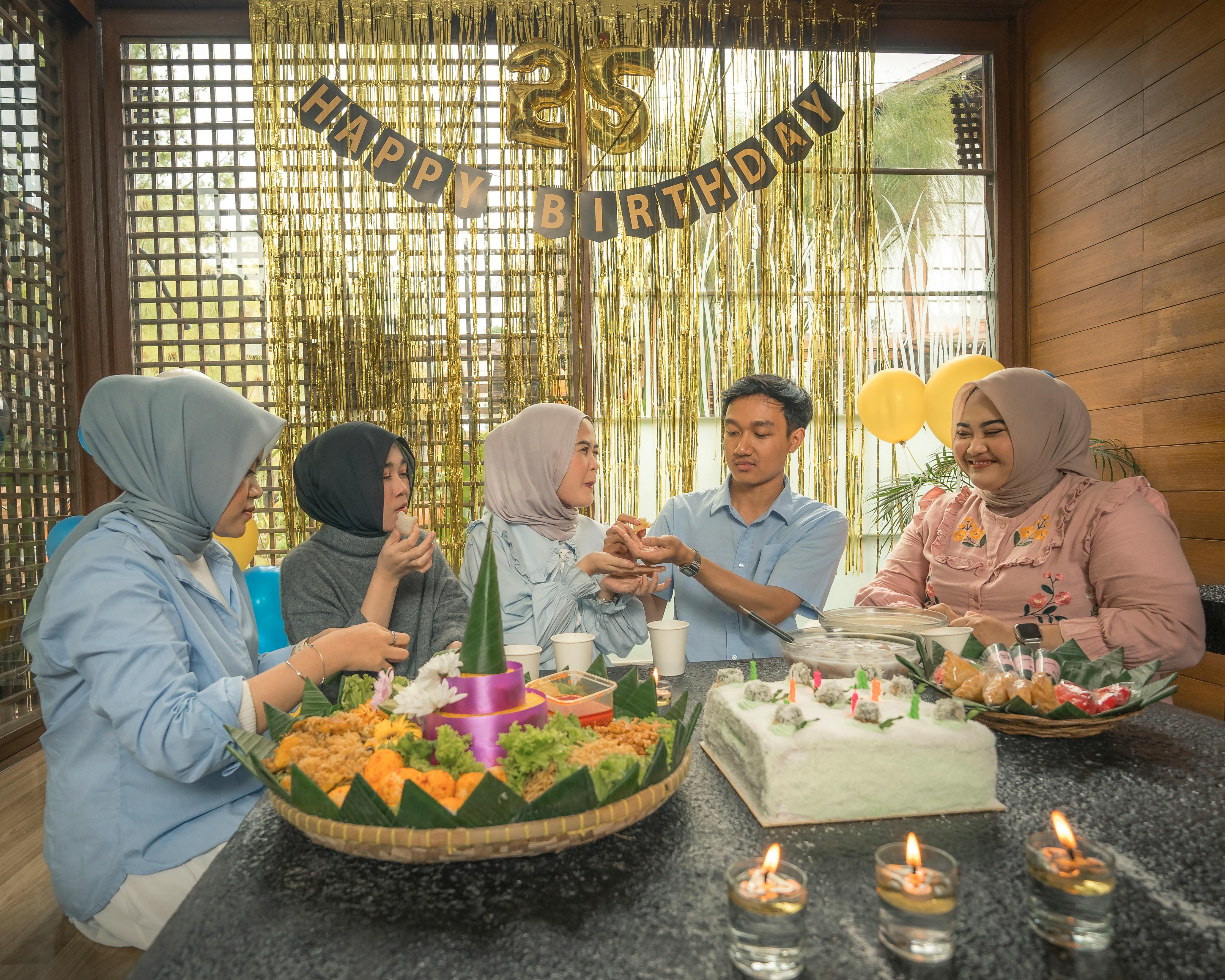 A group of friends celebrate a 25th birthday indoors with festive decorations and a traditional tumpeng dish.