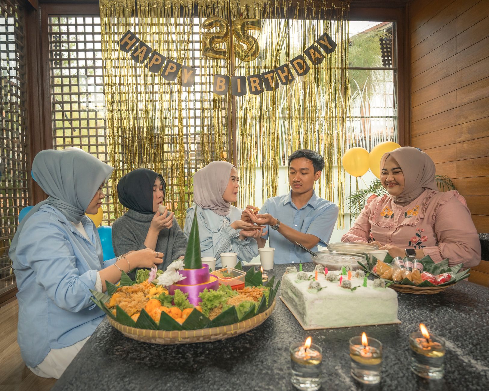 A group of friends celebrate a 25th birthday indoors with festive decorations and a traditional tumpeng dish.