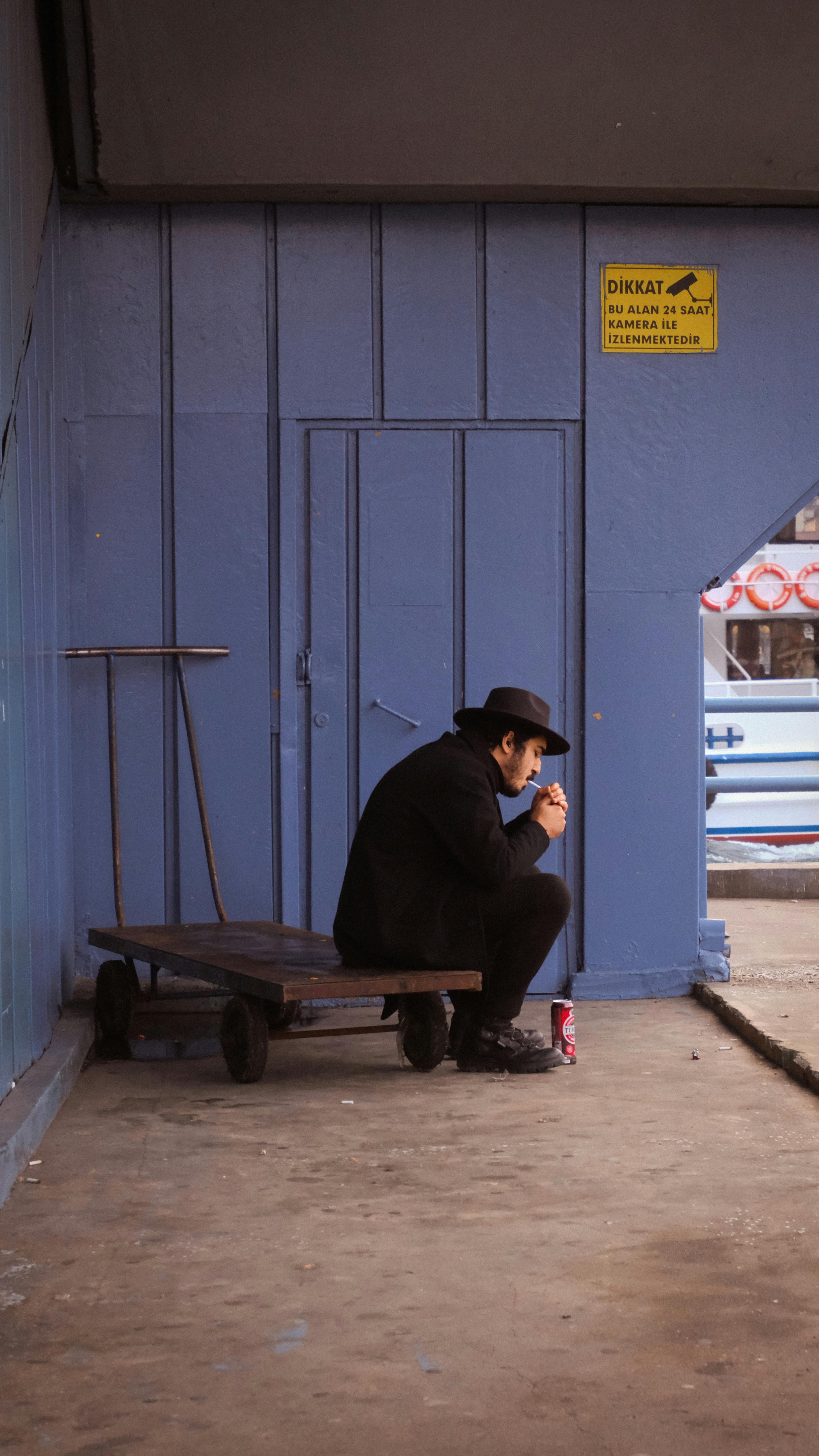 Free A contemplative man in a dark coat and hat sits on a trolley in a blue-painted indoor space. Stock Photo