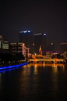 Vibrant cityscape with illuminated skyscrapers and bridge over calm river at night.