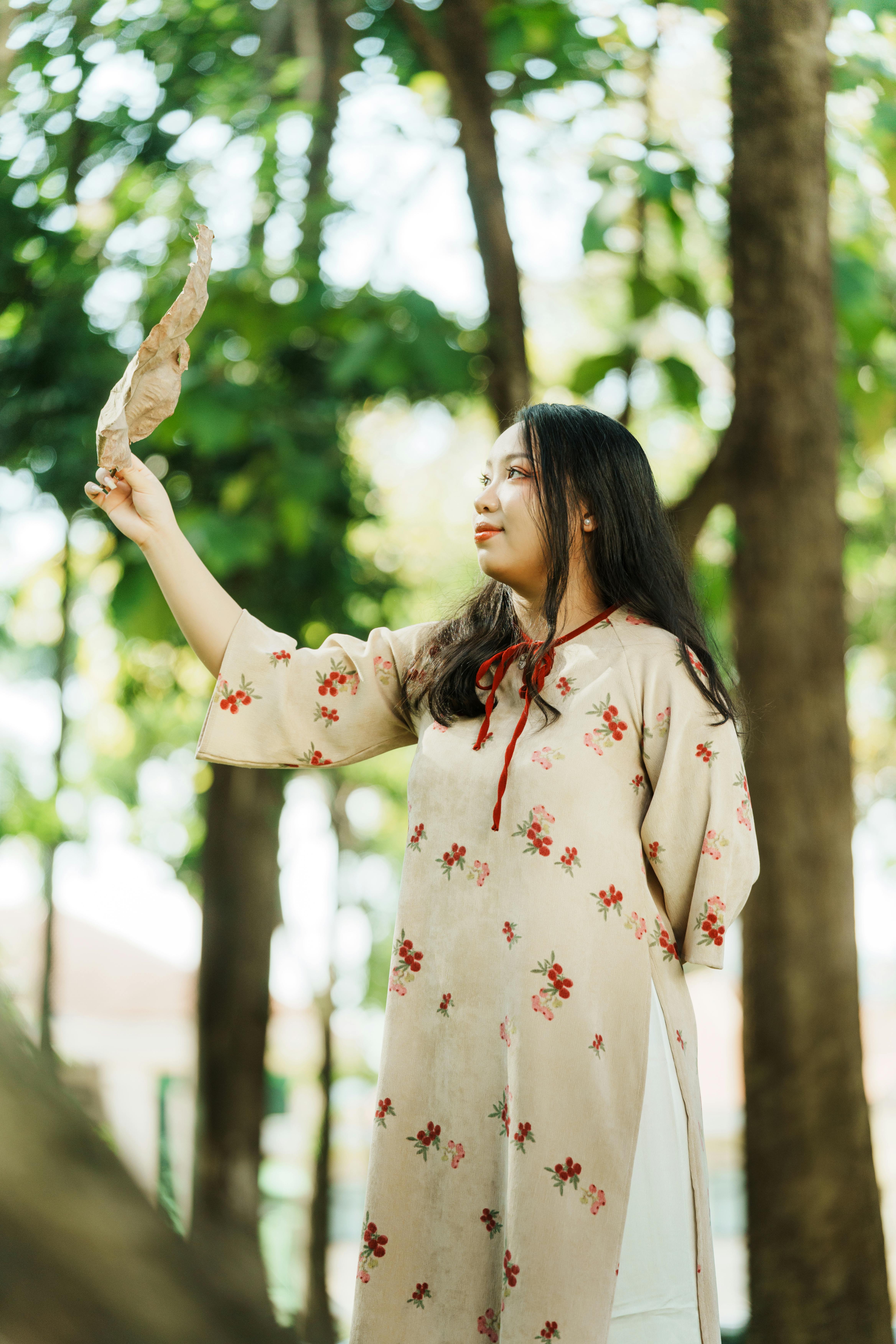 Gratis Una mujer joven con un vestido floral sostiene una hoja en medio de la exuberante vegetación de un bosque, capturando un momento sereno y contemplativo. Foto de stock
