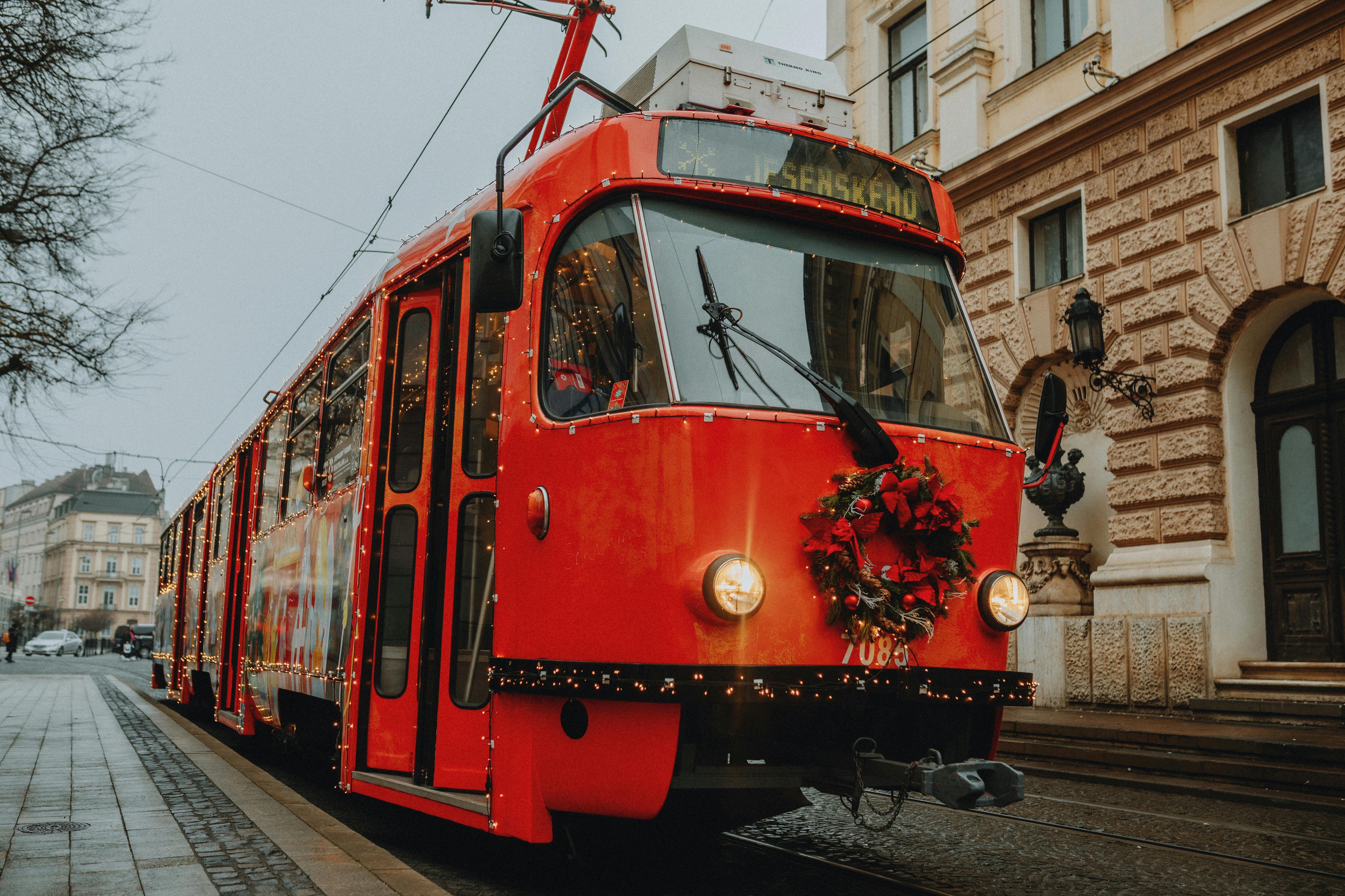 A vibrant red tram with festive holiday decorations travels through a city street lined with historic architecture.