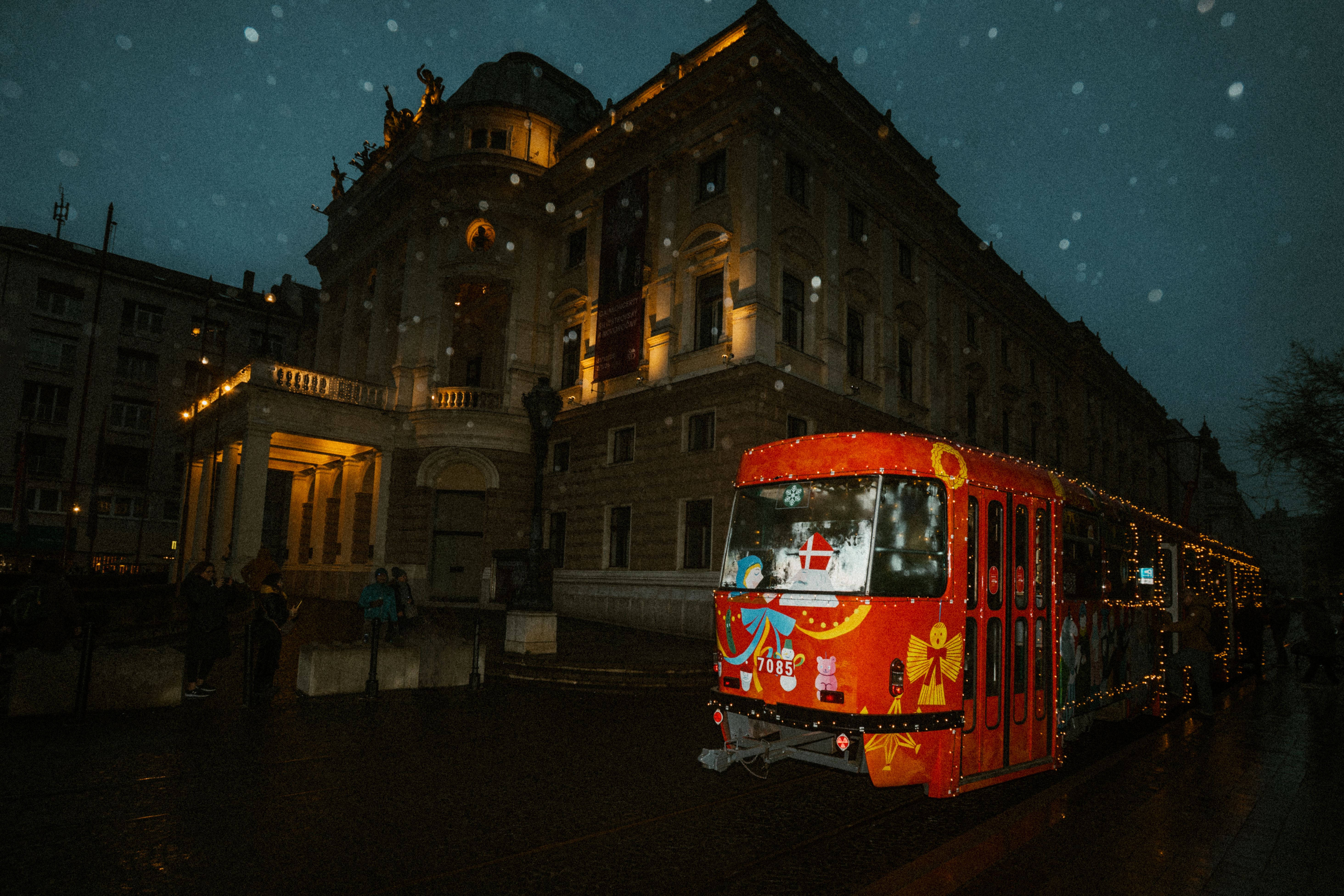 A decorated red tram on a snowy night by a historic building, creating a festive atmosphere.