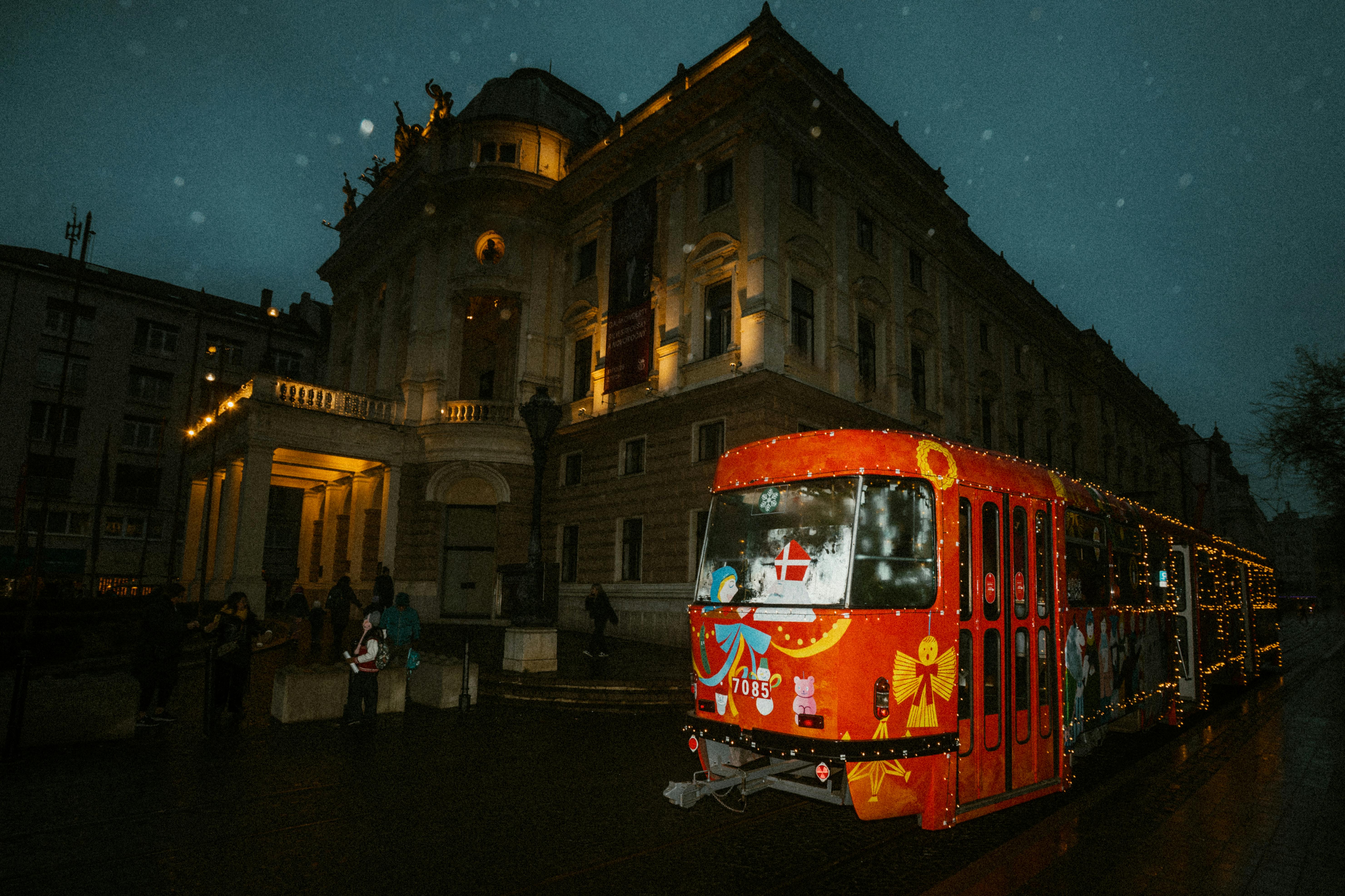 Brightly lit red tram adorned with festive lights passes in front of historical building at night.