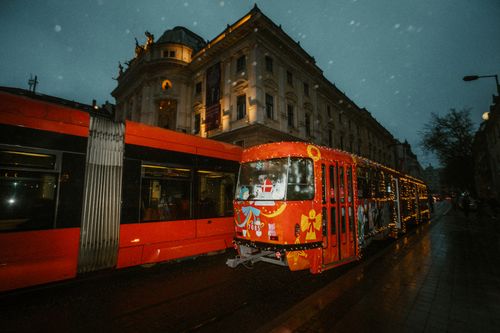 Free A decorated tram with Christmas lights travels past a historic building at dusk in a European city. Stock Photo