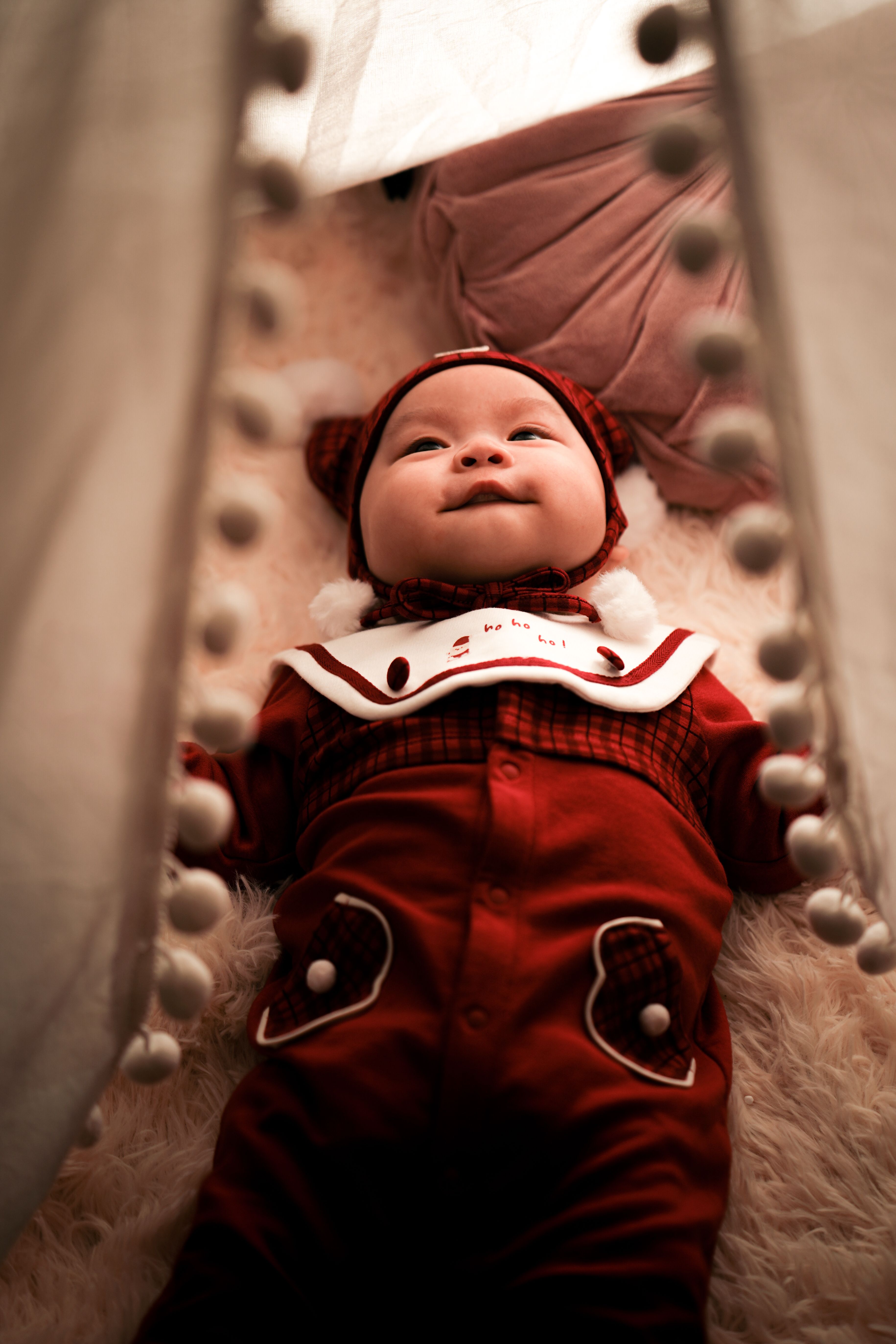 Cute baby dressed in red lies on a soft surface, framed by pom-pom curtain indoors.