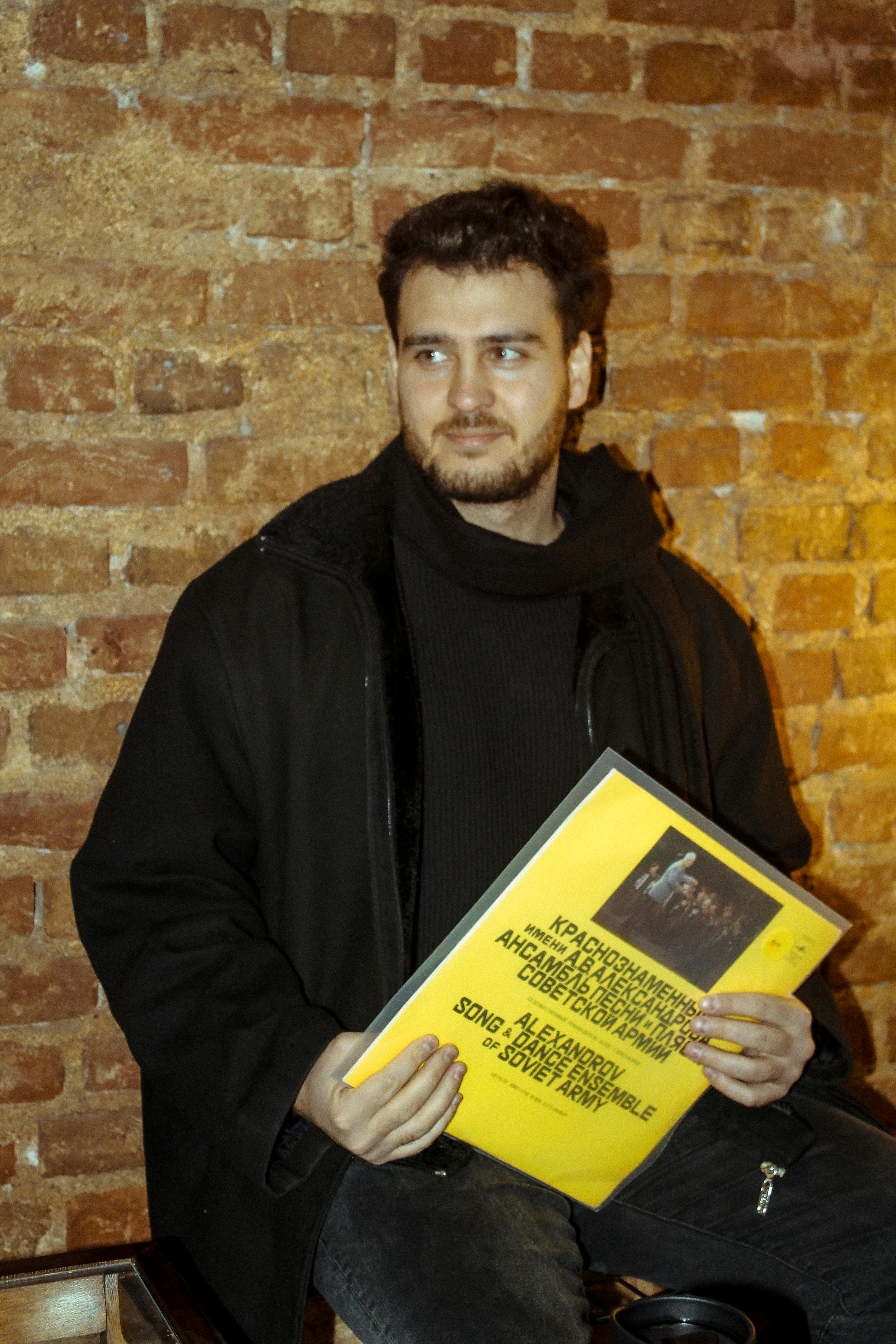Young man in black coat holding a yellow vinyl record, sitting against a brick wall.